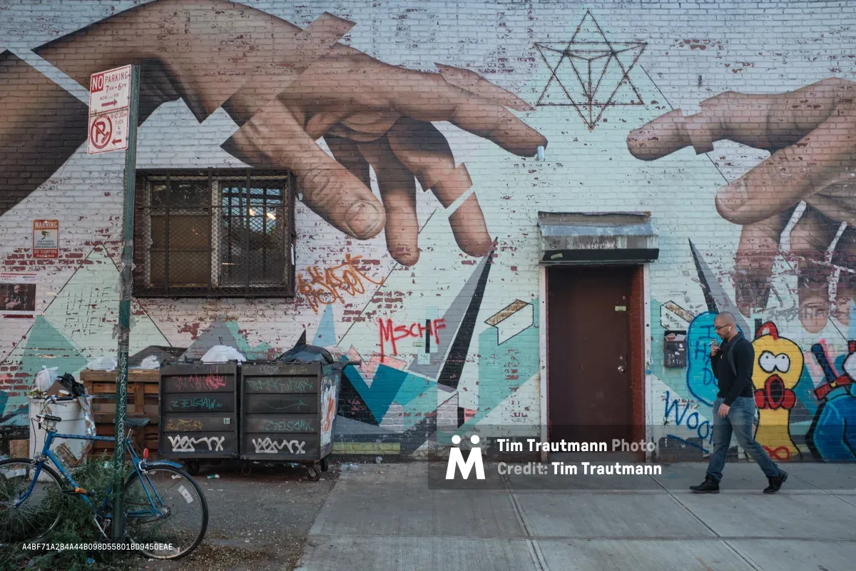 A lone figure in a black leather jacket strides past a vibrant mural on weathered brick walls in Williamsburg, Brooklyn. The street art features massive reaching hands, geometric patterns, and colorful character illustrations that transform the urban facade into a canvas of contemporary expression. A blue bicycle rests against dumpsters in the foreground, while peeling paint and layered graffiti tags speak to the neighborhood's authentic street culture. The overcast lighting creates a moody atmosphere that captures the raw creative energy of Brooklyn's artistic landscape.