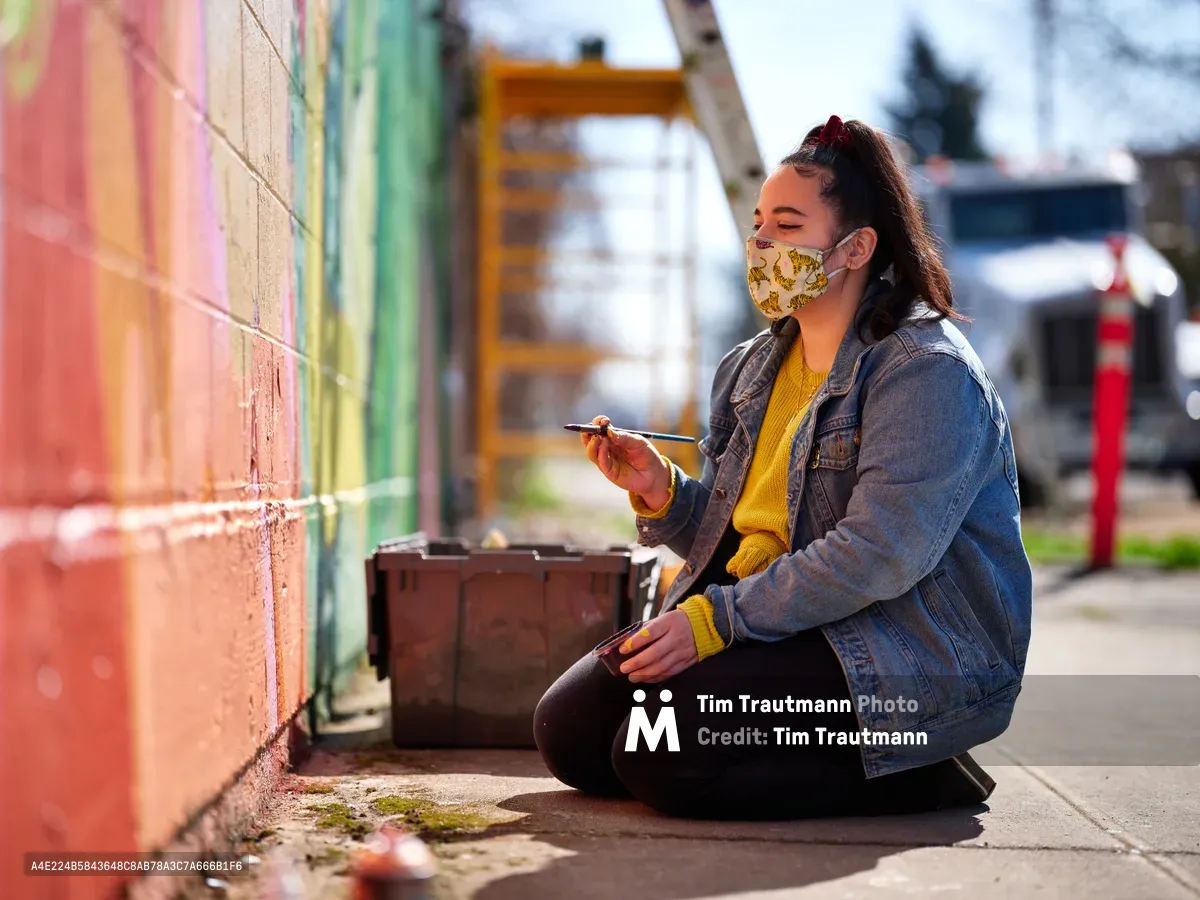 Multidisciplinary artist Vilatose sits cross-legged on the sidewalk, painting brush in hand, working on Jardín Cubana, a vibrant mural depicting three female figures along Alberta Street in Portland's historic arts district. Wearing a yellow patterned face mask and denim jacket over a mustard sweater, she pauses in her creative process against the colorful backdrop of pink, coral, and green paint strokes. Golden afternoon light bathes the scene, highlighting the weathered concrete and moss-touched pavement of this iconic Northeast Portland creative corridor.