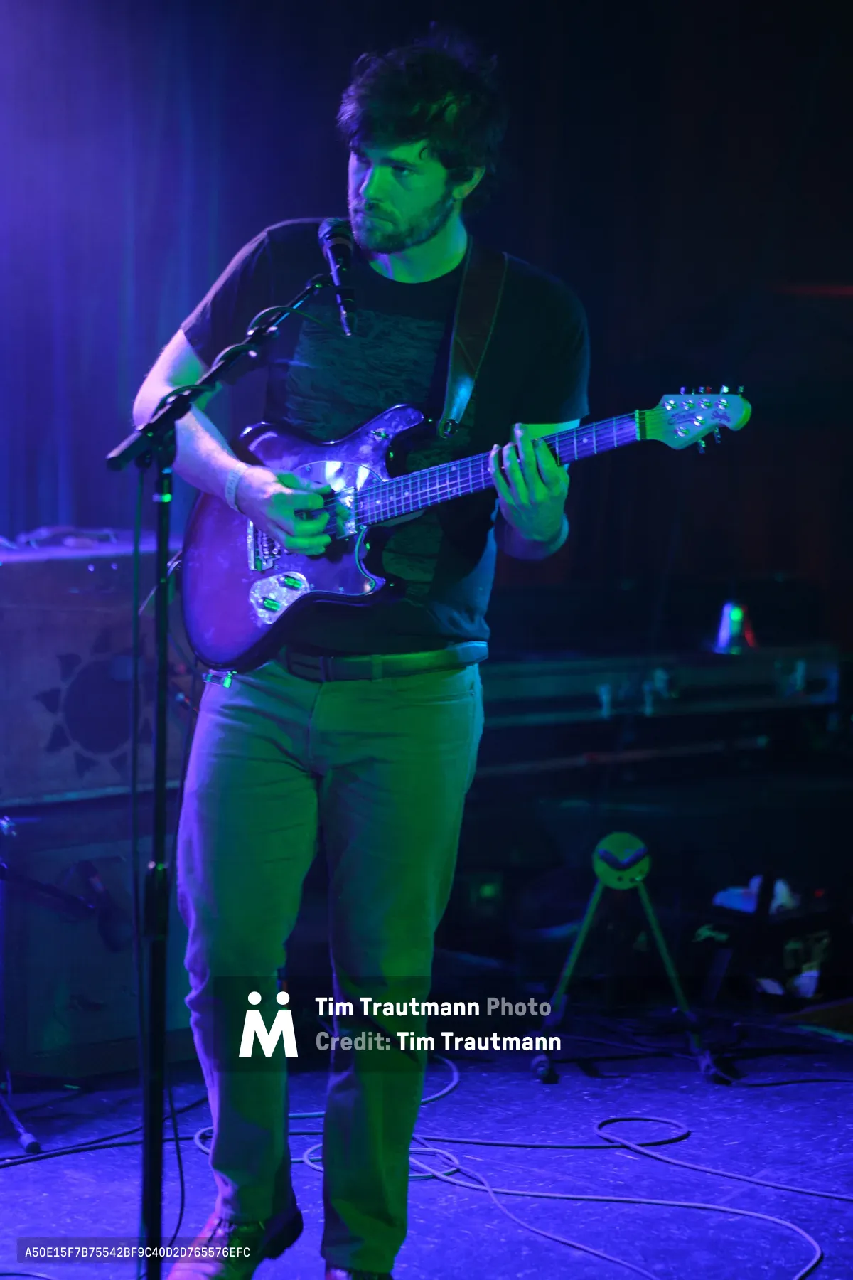 A bearded musician in a black t-shirt commands the intimate stage at Dante's in downtown Portland, his electric guitar caught mid-chord under saturated purple and green stage lighting. The atmospheric wash of color transforms the underground venue into a moody sanctuary where Sun Blood Stories weaves their sonic narrative. Cable-strewn floors and shadowed equipment create texture in the foreground while the performer's concentrated expression speaks to the focused intensity of this experimental band's mesmerizing live sound. Originally from Boise, Idaho and now Portland-based, the group channels their operatic approach to music through every chord.