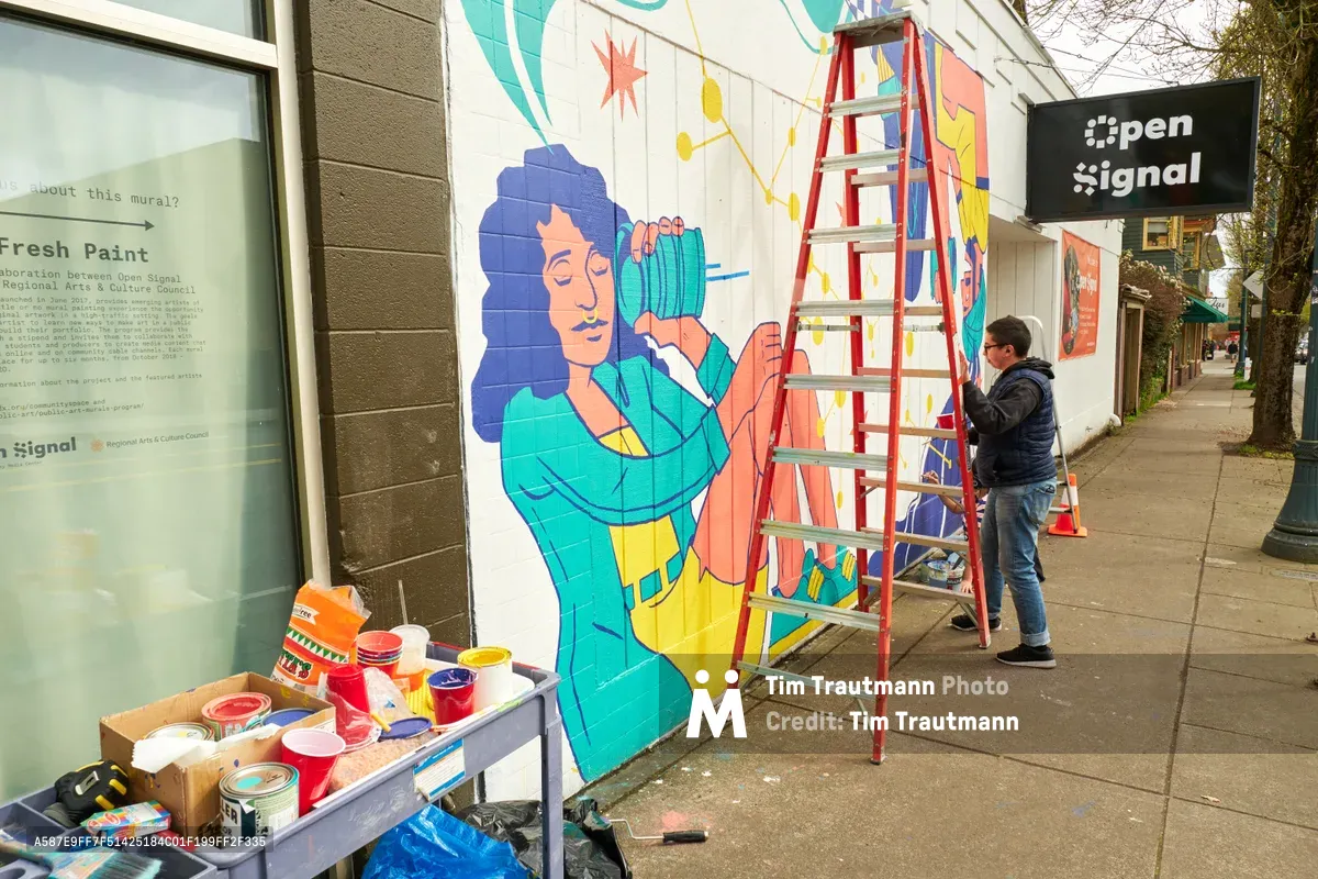 Artist Anke Gladnick works on a vibrant community mural titled "Let's Talk" on the exterior wall of Open Signal in Portland's Eliot neighborhood. The colorful artwork depicts a figure with blue hair holding what appears to be a vintage camera or recording device, rendered in bright teals, yellows, and coral tones. A red stepladder stands against the wall as the artist adds finishing touches, while paint cans and supplies are organized on a nearby table, capturing the collaborative spirit of public art creation.