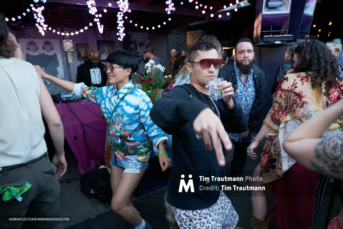 Revelers lose themselves in the rhythm at Your Sunday Best day party, bathed in the moody purple glow of string lights cascading from White Owl Social Club's ceiling. A woman in a vibrant tropical print shirt dances with abandon while a man in sunglasses and black clothing extends his hand toward the camera, creating an intimate connection with the viewer. The crowded dance floor pulses with eclectic fashion choices and uninhibited movement, capturing the essence of Portland's underground party culture in this dimly lit, art-adorned venue.