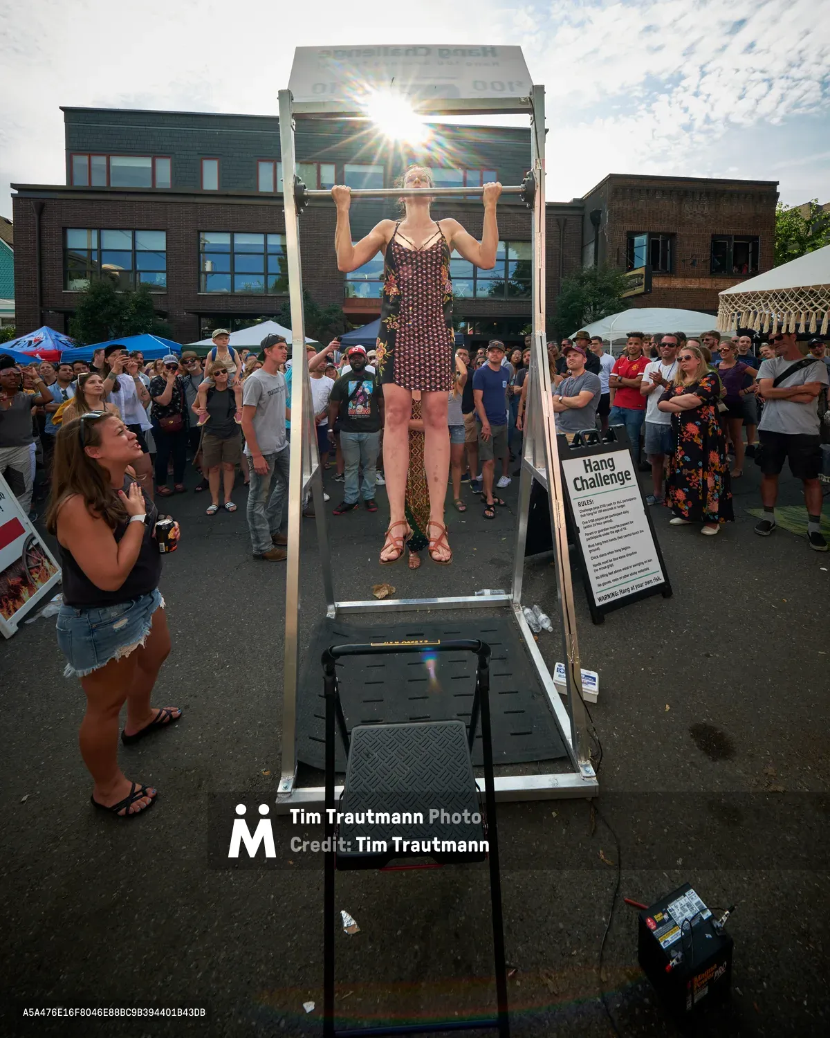 A determined woman in a geometric patterned dress executes a perfect dead hang from a pull-up bar at Portland's Mississippi Street Fair, her form silhouetted against the brilliant afternoon sun. The crowd of onlookers forms a semicircle around the Hang Challenge station, their faces a mixture of encouragement and awe as she demonstrates raw strength in an unexpected urban setting. The lens flare creates dramatic rays that pierce through the scene, while the contemporary brick architecture of North Mississippi Avenue provides a distinctly Portland backdrop to this moment of athletic determination.