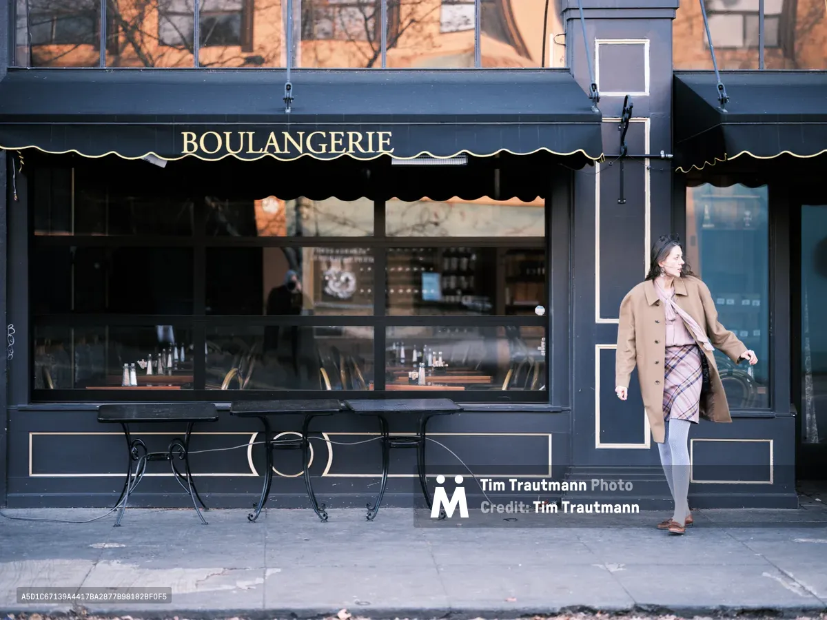 A woman in a camel coat and plaid skirt walks past a charming French boulangerie on Portland's Northeast Alberta Street. Golden morning light washes the scene, creating a warm glow against the dark navy storefront with its elegant gilt lettering. The intimate street-level composition captures the quiet rhythm of neighborhood life, with wrought iron cafe tables awaiting the day's first customers.