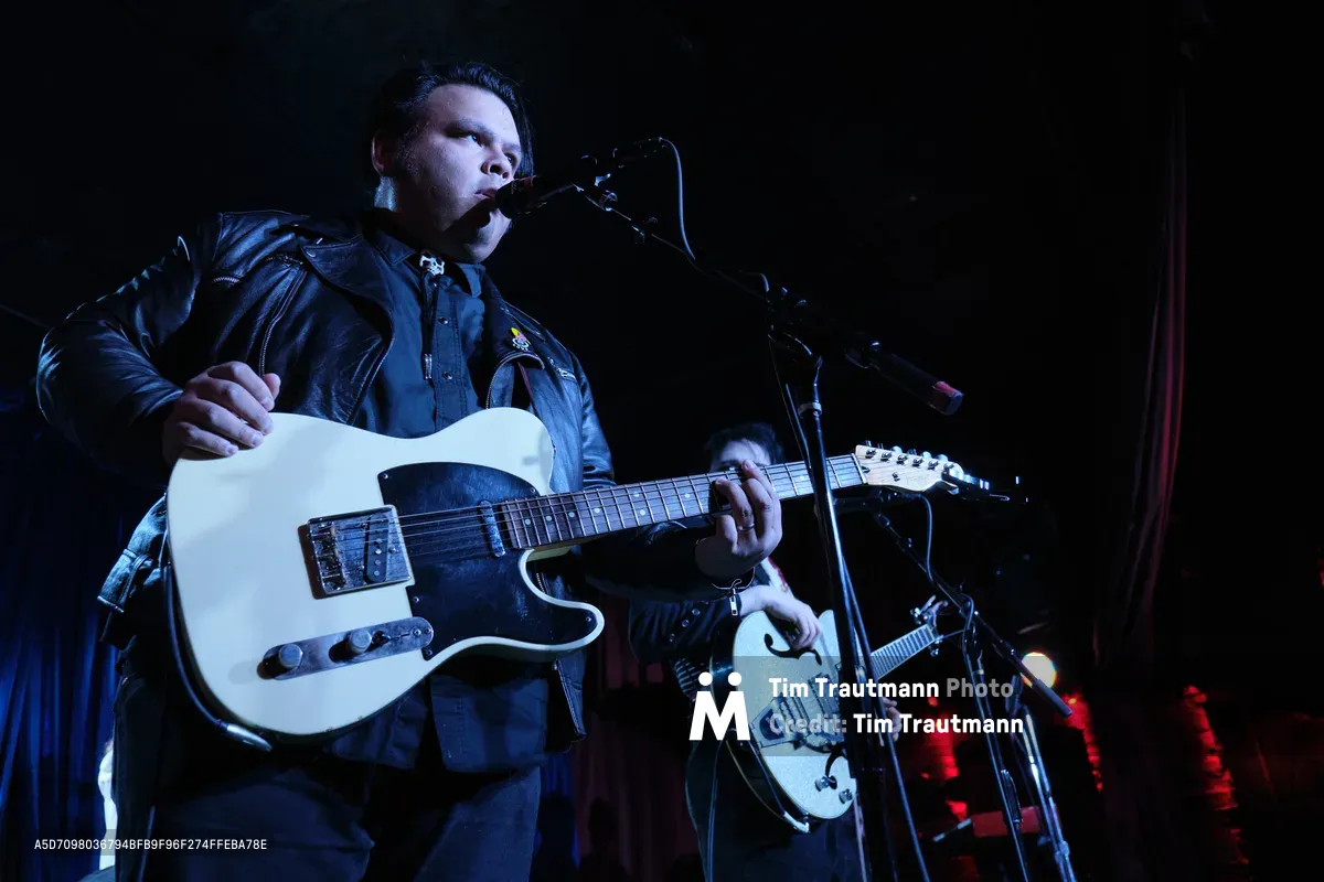 Victor Franco of Roselit Bone commands the shadowed stage at Dante's, his white Telecaster gleaming against the venue's inky atmosphere. Bathed in moody blue stage lighting with crimson accents bleeding from the background, Franco leans into the microphone with focused intensity while his bandmate's hollow-body guitar catches fragments of light in the periphery. The intimate Portland club setting creates a cocoon of sound and shadow, where the performer's leather jacket and concentrated expression speak to the raw authenticity of the underground music scene.