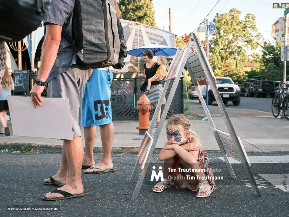 A young girl with a blue butterfly face painting sits curled up on the street beneath a metal A-frame ladder sign, hugging her knees with a sullen expression at the Last Thursday street fair on Alberta Street in Portland, Oregon. She wears a floral dress and white sandals. The legs of two adults standing nearby are visible in the foreground, and a woman holding a toddler is visible in the background near a blue patio umbrella and a red fire hydrant.
