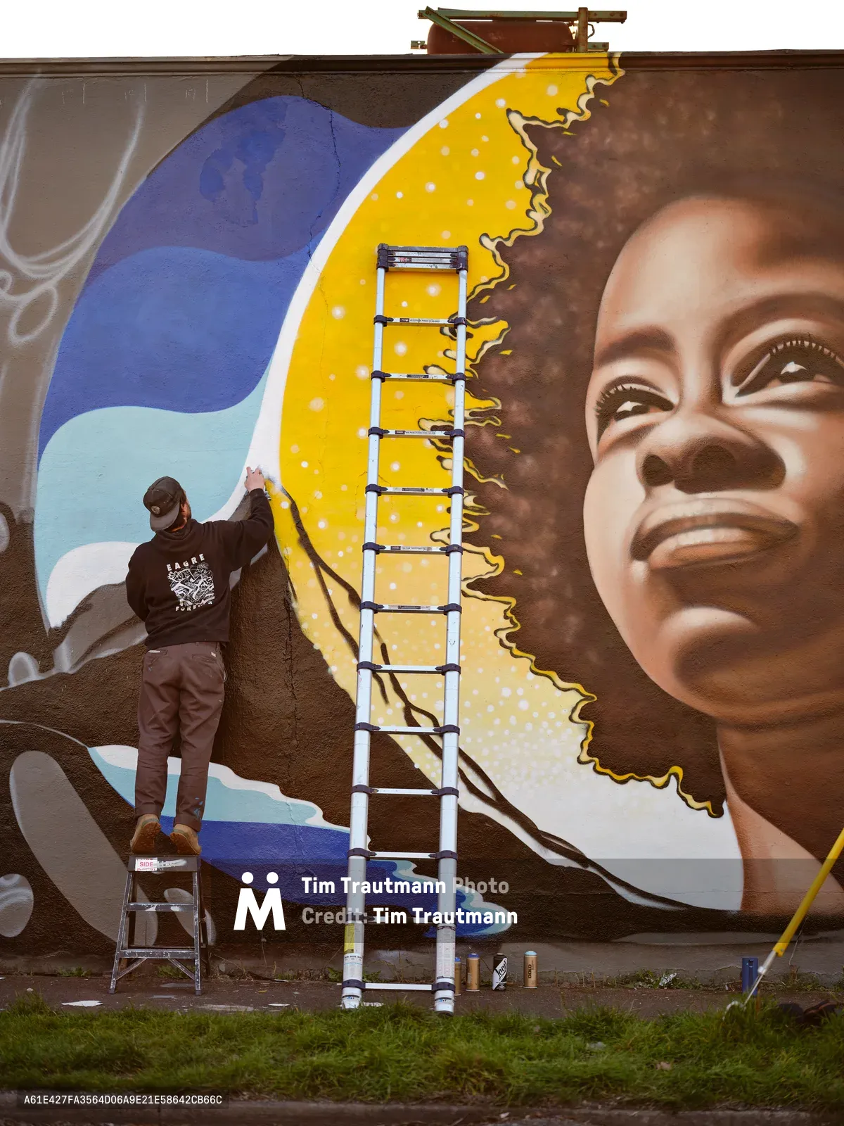 Street artist Travis Fields, working under the name campographic, applies vibrant paint to a towering mural of poet Amanda Gorman on a brick wall in Portland's Alberta district. The artwork depicts Gorman with an upward gaze against a brilliant yellow sun and flowing blue waves, while Fields balances on a stepladder, his dark hoodie contrasting against the warm tones of his creation. An aluminum extension ladder stretches upward along the wall, with paint cans scattered on the grass below, capturing the active process of public art creation in this Northeast Portland neighborhood.