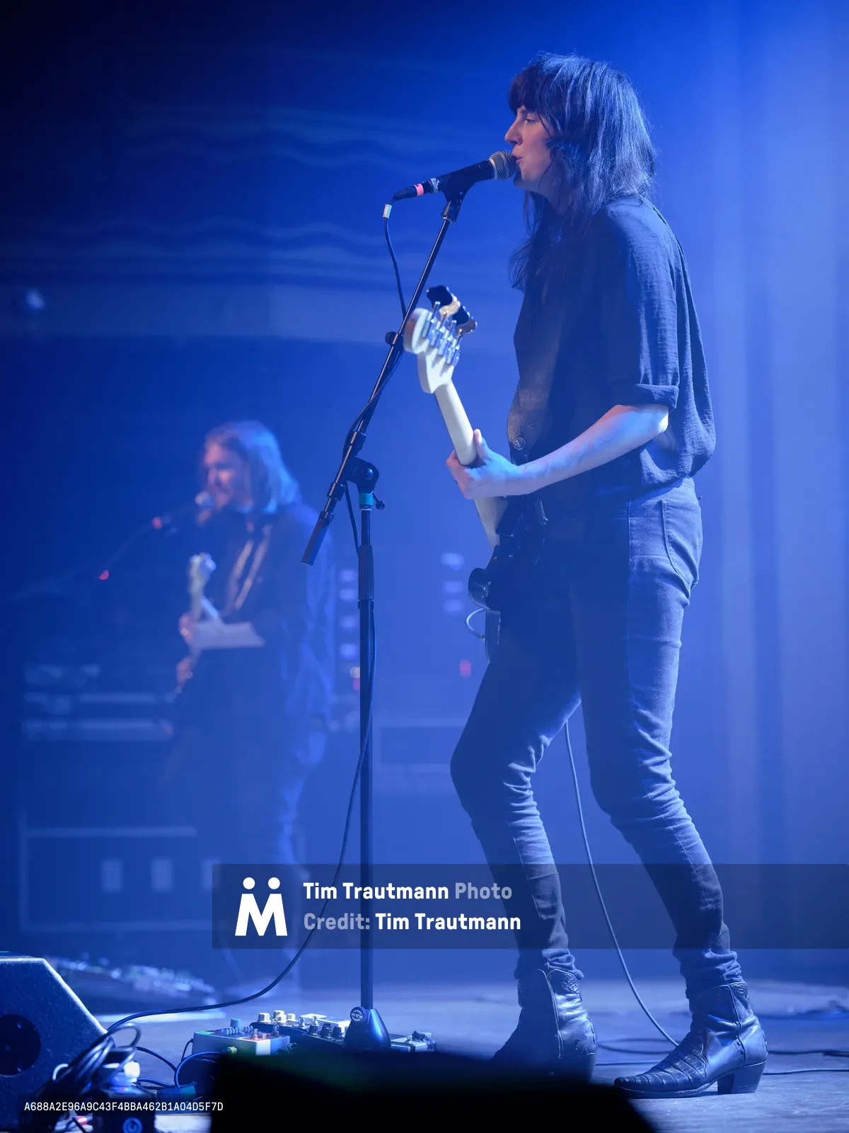 Emma Richardson of Band of Skulls performs under saturated blue stage lighting at Webster Hall in Manhattan, her silhouette defined against the moody atmospheric glow. The intimate venue setting captures the raw energy of the anniversary tour, with Richardson positioned at center stage while bandmate Russell Marsden remains partially visible in the background shadows. Her focused expression and commanding stage presence anchor this moment of live musical performance in the iconic East Village venue.