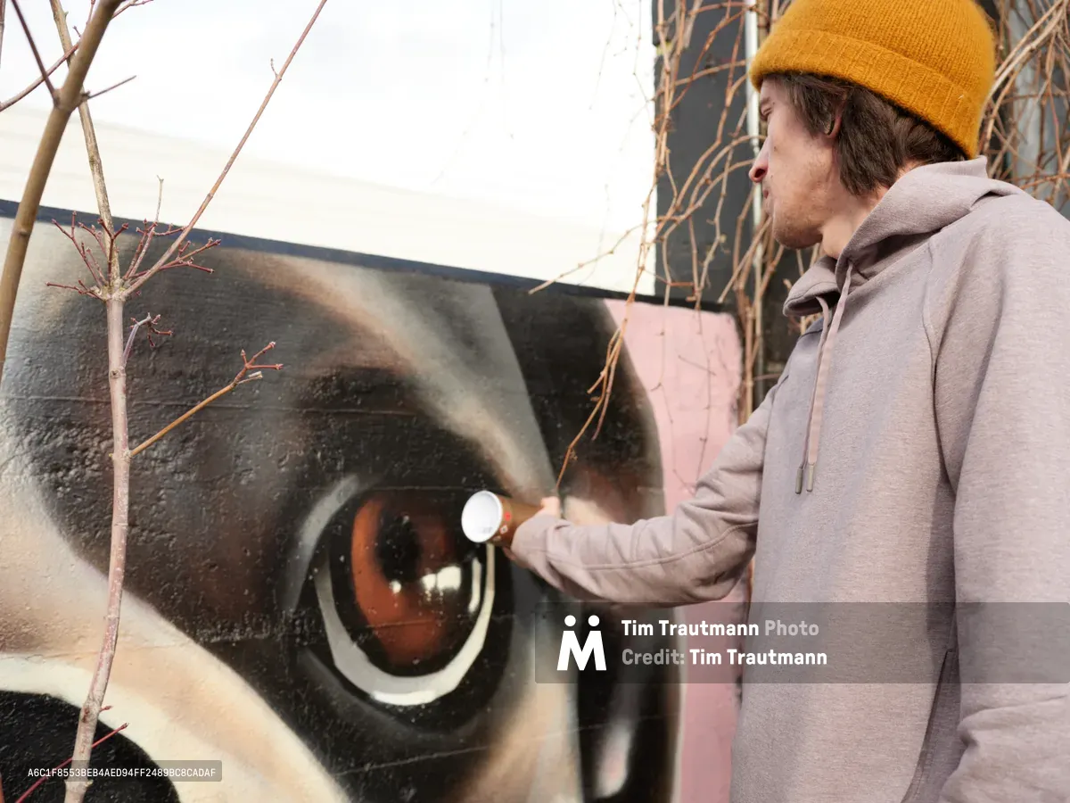 Muralist Wetiko applies spray paint to a captivating hyperrealistic eye mural on an exterior wall in Portland's Hosford-Abernethy neighborhood. Dressed in a mustard yellow beanie and gray hoodie, the artist works with focused concentration as golden hour light filters through bare winter branches, casting dramatic shadows across the detailed amber iris. The intimate documentary moment captures the transformative power of street art in the Clay Creative district.