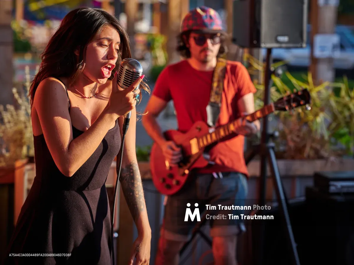 Aubrey Gordon commands the microphone with passionate intensity during a golden hour performance at Portland Mercado in Southeast Portland. The warm, amber light bathes the vocalist in a nostalgic glow as she delivers her set, while her guitarist provides atmospheric accompaniment in the softly focused background. The intimate outdoor venue, with its colorful murals and lush greenery, creates a quintessentially Portland music scene that captures the city's vibrant cultural spirit.