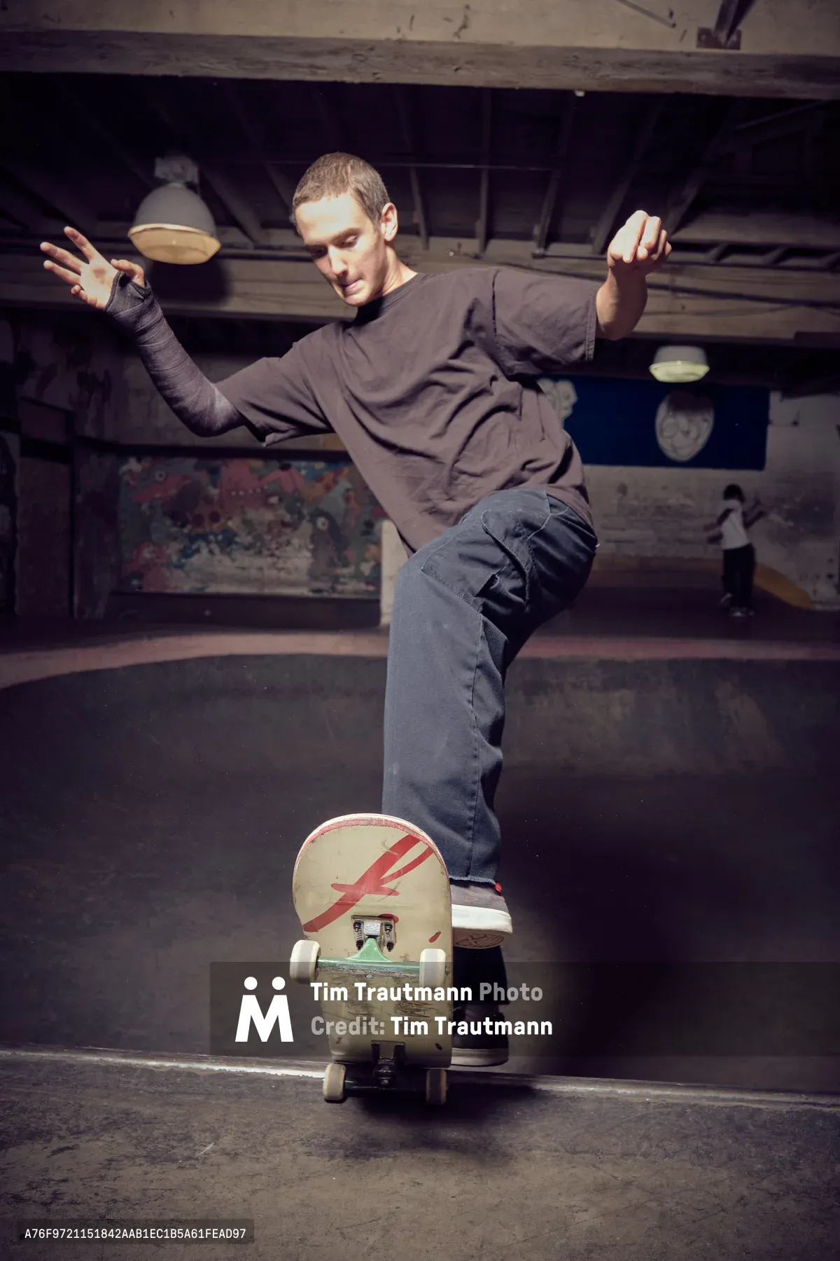 A young skater demonstrates perfect balance while riding a distinctive red lightning bolt-decorated skateboard through the gritty interior of Commonwealth Skateboarding in Portland's Buckman neighborhood. His arms extend gracefully for stability as he navigates the worn concrete floors beneath exposed ceiling beams and industrial lighting. The atmospheric shot captures the raw authenticity of skate culture within this iconic Southeast Portland skate shop, where weathered surfaces and graffitied walls create a backdrop steeped in street credibility.