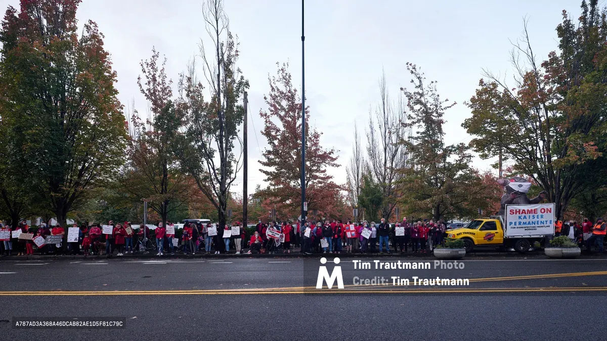 A sea of red-clad nurses and supporters mass along Northeast Multnomah Street outside Kaiser Permanente Tower in Portland's Lloyd District, their protest signs creating a rhythmic pattern against autumn's burnished foliage. The overcast Pacific Northwest sky casts diffused light across the demonstration, while a yellow union truck bearing "SHAME ON KAISER PERMANENTE" anchors the right side of the frame. The striking healthcare workers stretch in an unbroken line along the sidewalk, their collective presence transforming the corporate streetscape into a theater of labor solidarity.