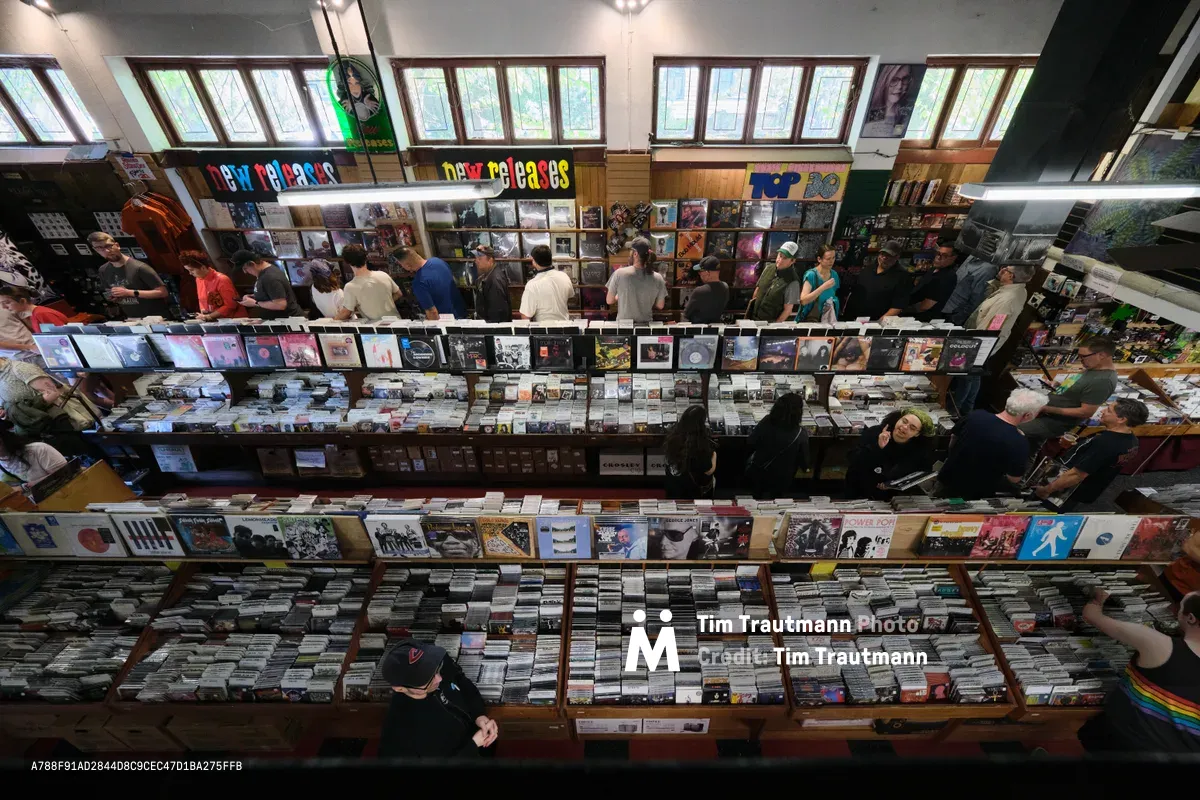 A bird's-eye view captures the bustling atmosphere of Record Store Day 2026 at Music Millennium on East Burnside Street in Portland, Oregon. Dozens of vinyl enthusiasts browse through methodically organized rows of records, their silhouettes creating patterns against the warm wooden flooring. Natural light streams through tall windows along the storefront, illuminating colorful album covers and handwritten signage that celebrates the enduring ritual of analog music discovery.