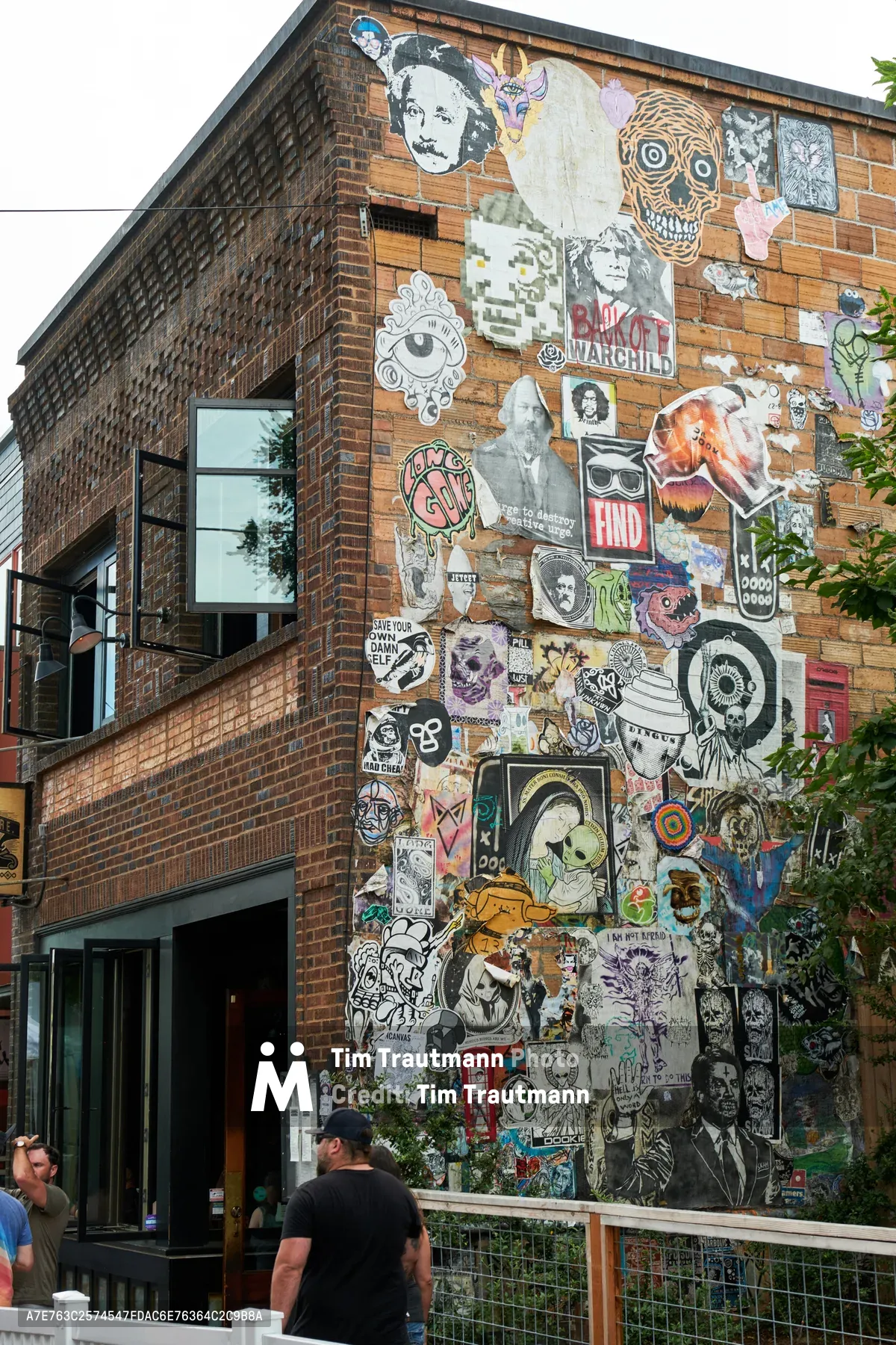 A vibrant corner building in Portland's Mississippi District showcases an eclectic collage of street art, stickers, and wheatpaste covering its weathered brick facade. The layered artwork creates a chaotic tapestry of pop culture references, portraits, skulls, and graphic designs that speak to the neighborhood's creative underground culture. Two pedestrians in casual clothing walk past the decorated corner, while modern storefront windows reflect the overcast daylight typical of Oregon's urban landscape.
