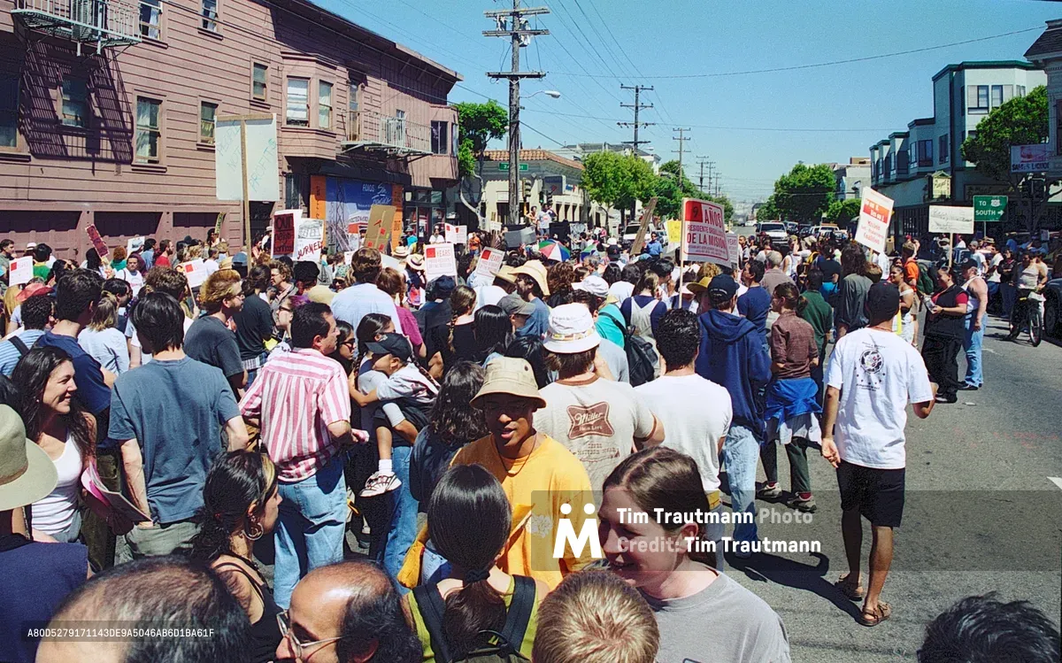 Hundreds of residents flood Bryant Street in San Francisco's Mission District during a passionate anti-displacement demonstration organized by the Mission Anti-Displacement Coalition. The diverse crowd carries protest signs advocating for affordable housing and community preservation under bright afternoon sunlight. Victorian-era buildings and utility poles frame the residential street scene as neighbors unite against gentrification pressures threatening the neighborhood's working-class and artistic communities.