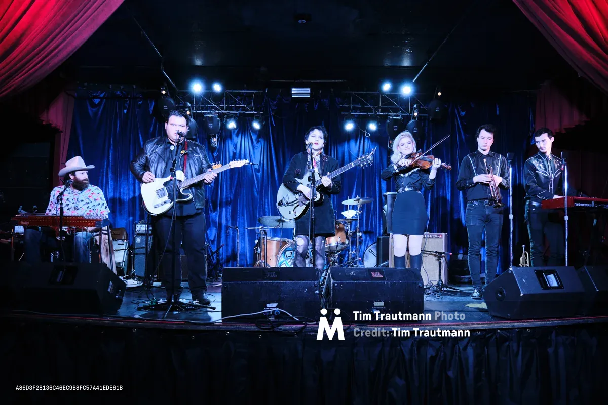Six musicians of Roselit Bone prepare for their performance during a pre-show soundcheck at the intimate Dante's venue in Portland, Oregon. The band assembles on stage beneath dramatic blue and red theatrical lighting, with instruments including electric guitars, violin, and keyboards creating an atmospheric tableau. Crimson curtains frame the scene while professional stage lights pierce through the venue's characteristic darkness, capturing the focused energy of rehearsal at this legendary Portland music club.