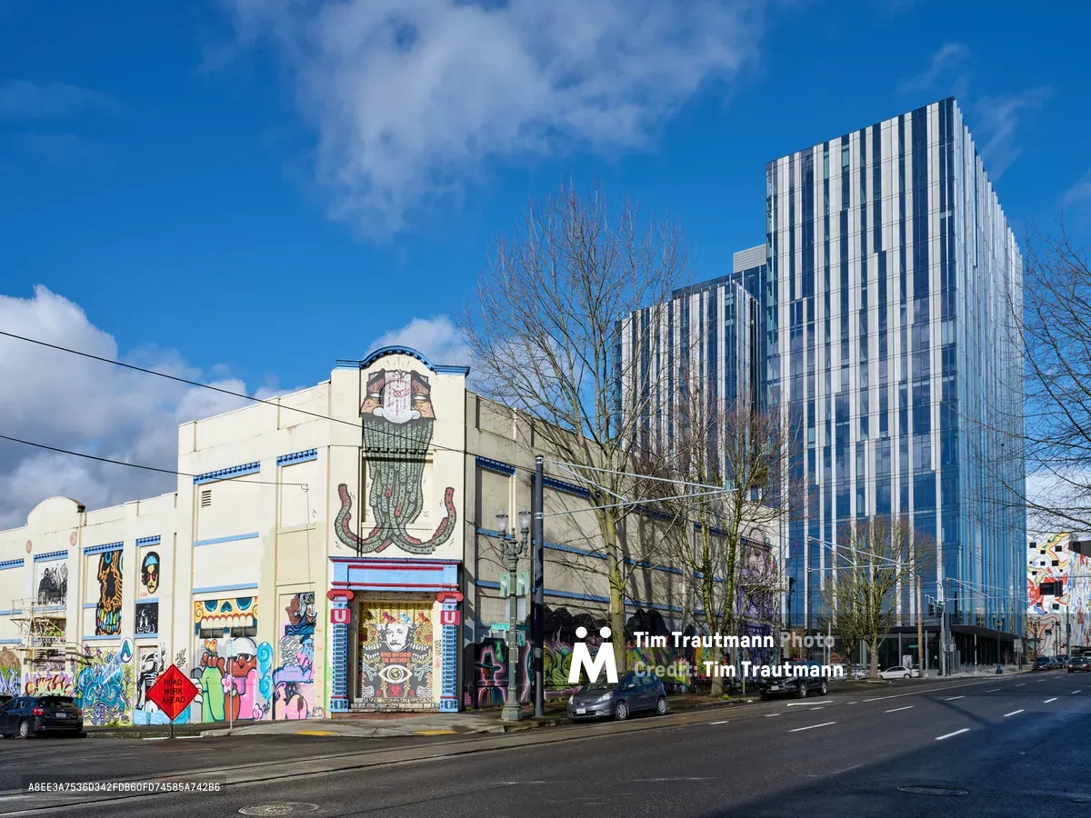 A vibrant collision of architectural eras unfolds along Southeast Ash Street in Portland's Central Eastside, where the weathered concrete facade of a former Salvation Army warehouse blooms with commissioned street murals. The building's utilitarian bones, adorned with colorful graffiti art featuring cartoon characters, abstract forms, and eye-catching imagery, stand in stark contrast to the gleaming glass towers rising behind it. Winter-bare trees frame the scene under a brilliant blue sky punctuated by white clouds, while parked cars line the quiet urban street, capturing the dynamic tension between Portland's industrial past and its contemporary creative renaissance.