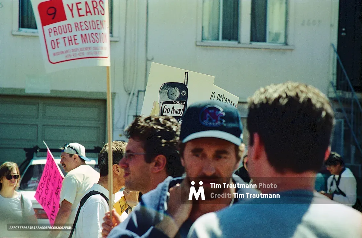 Sunlight filters through the crowd as Mission District residents gather for an anti-displacement demonstration on Bryant Street. A man in a baseball cap stands near a handmade sign reading 'NOKLU Go Away'—a clever play on Nokia referencing tech worker culture—while another protester displays '9 Years Proud Resident of The Mission' on a red placard. The intimate street scene captures the grassroots energy of longtime residents fighting gentrification, with weathered Victorian architecture providing backdrop to this community activism moment.