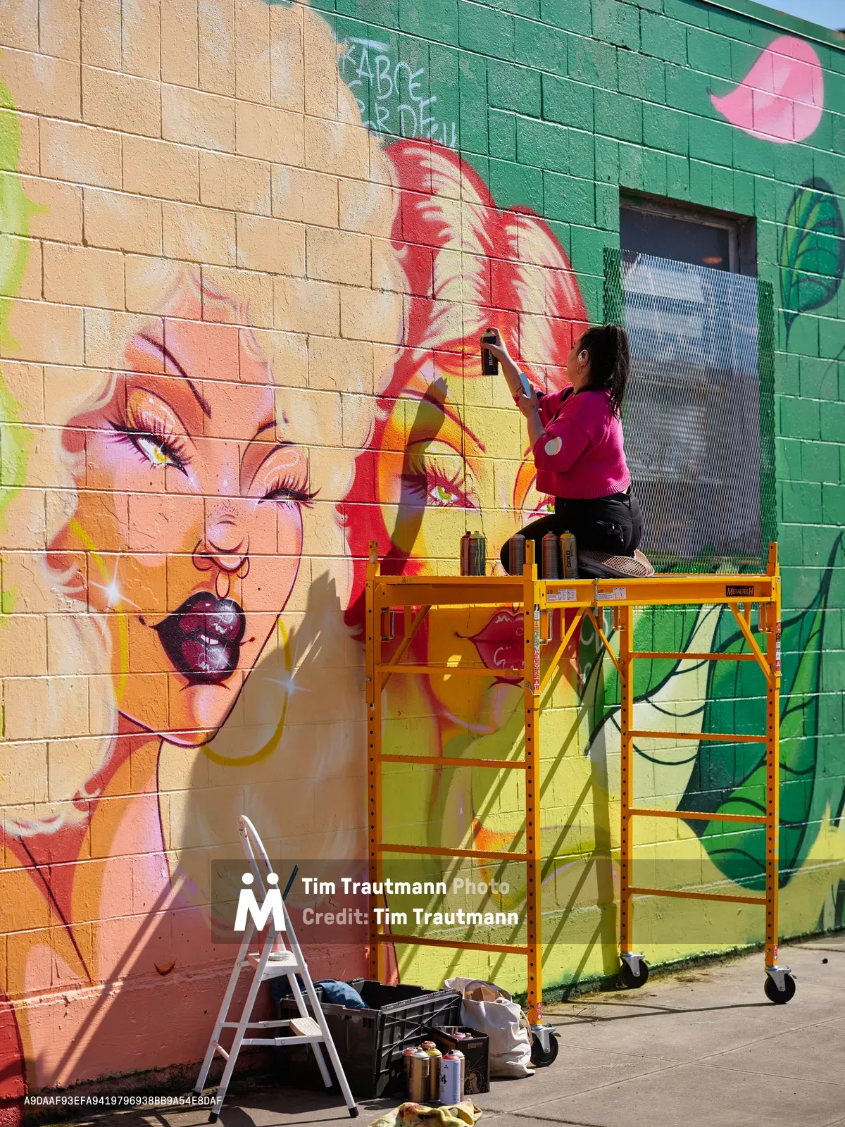 Multidisciplinary artist Vilatose works from an orange scaffolding platform, adding vibrant details to a large-scale mural on Northeast Alberta Street in Portland's Alberta Arts District. The colorful artwork features a portrait of a woman with flowing red hair and dramatic makeup against a backdrop of warm peach and emerald green blocks. Afternoon sunlight casts sharp shadows across the concrete sidewalk, where paint supplies and a step ladder await below the industrial mobile scaffold.