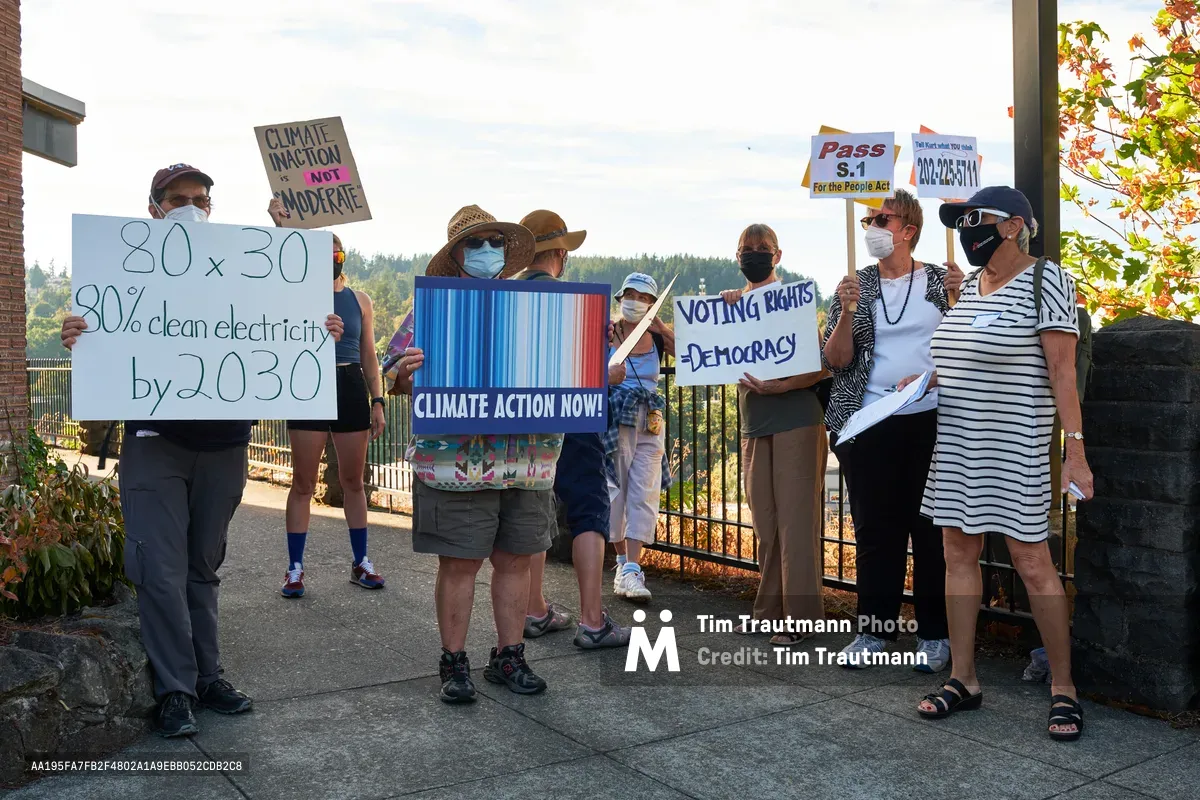 A diverse group of masked protesters gathers on the sidewalk outside Representative Kurt Schrader's office in Oregon City, holding handwritten signs advocating for clean energy and climate action. The late afternoon light bathes the scene in warm tones as demonstrators display messages calling for 80x30 clean electricity goals, voting rights, and urgent climate action. Autumn foliage frames the peaceful assembly, while brick buildings and iron fencing provide an urban governmental backdrop to this grassroots political demonstration.
