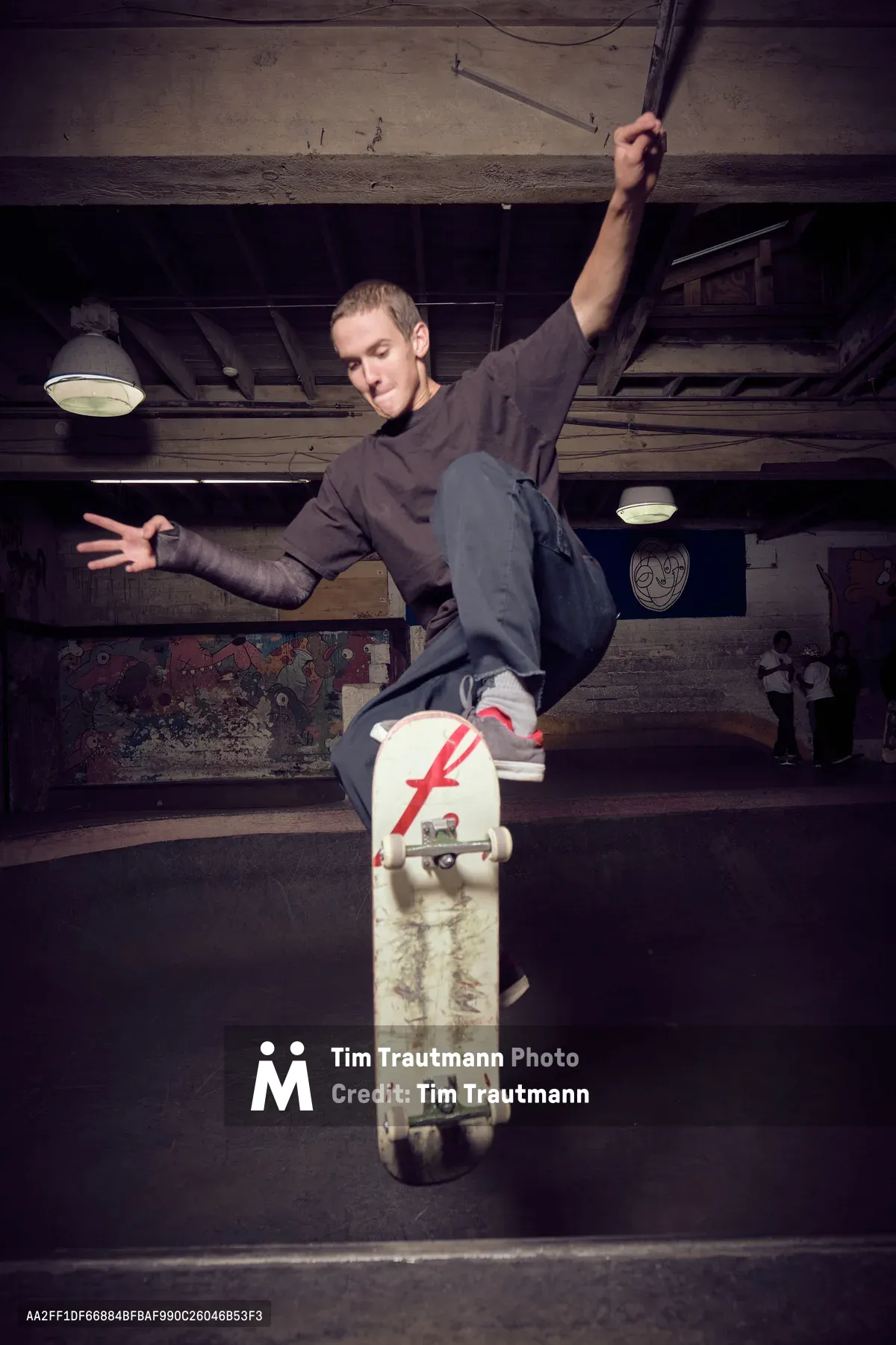 A young skateboarder launches into motion beneath the weathered wooden beams of Commonwealth Skateboarding's subterranean skate space in Portland's Buckman neighborhood. Illuminated by industrial pendant lights, the athlete grips his white skateboard marked with a distinctive red 'F' logo while suspended mid-trick against the basement's rustic architecture. The raw concrete ceiling and exposed timber framework create a gritty urban sanctuary where street skating culture thrives in the shadows.