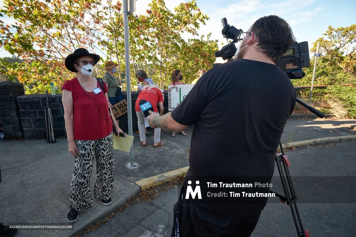 A masked climate activist in a burgundy top and patterned pants stands confidently before a television camera crew in Oregon City's McLoughlin Promenade. The woman, wearing a black hat and sunglasses, speaks into a KOIN Channel 6 microphone while fellow protesters gather in the background beneath autumn foliage. Golden afternoon light filters through the trees, casting long shadows on the concrete sidewalk where this grassroots demonstration unfolds outside Representative Kurt Schrader's office.