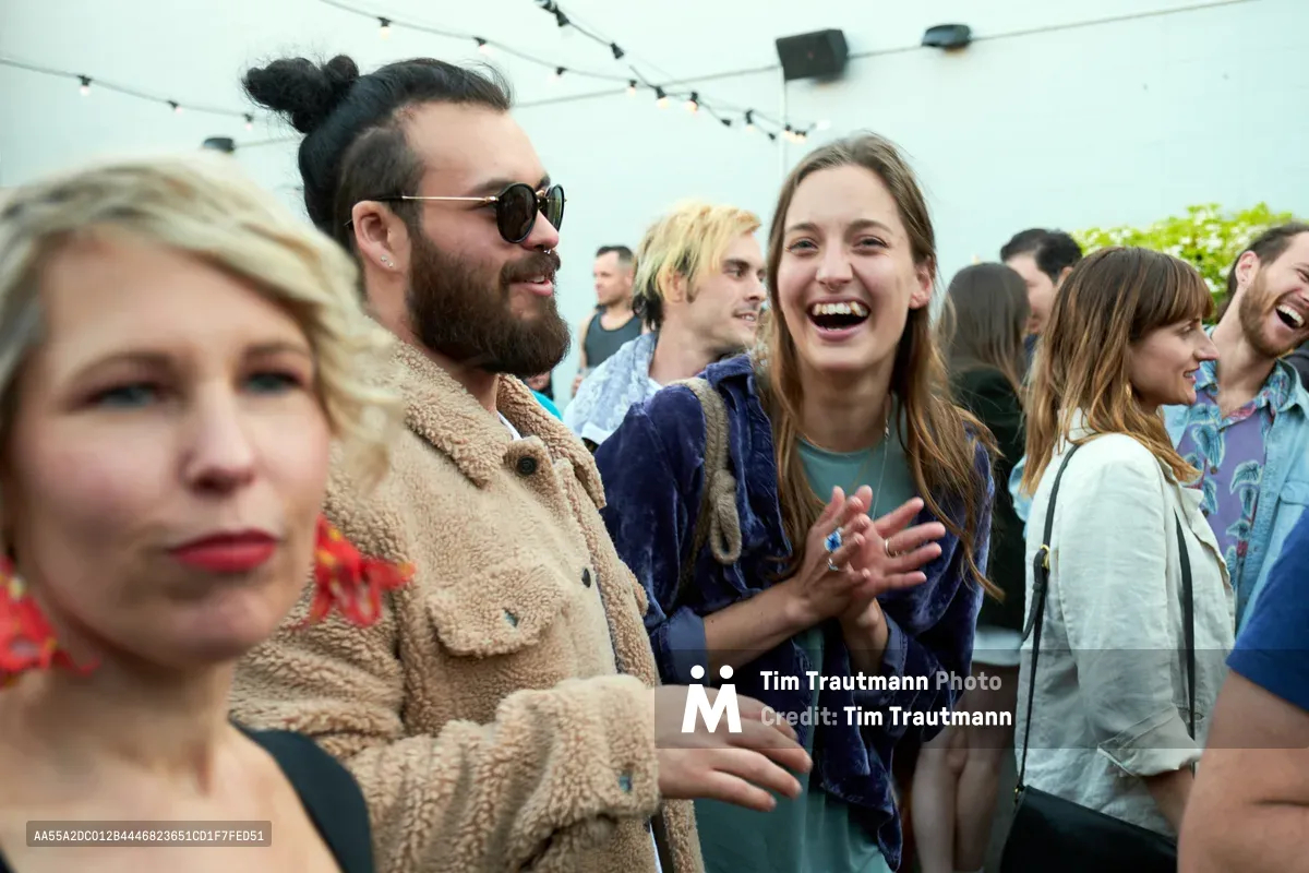 Under the soft glow of string lights at Portland's White Owl Social Club, a diverse crowd of young adults celebrates at Your Sunday Best's season opener. A bearded man in sunglasses and sherpa jacket shares an animated moment with a laughing brunette woman in navy velvet, while a blonde woman in a textured coat occupies the foreground. The outdoor patio buzzes with the energy of summer socializing, capturing the spontaneous joy of Portland's vibrant day party scene.