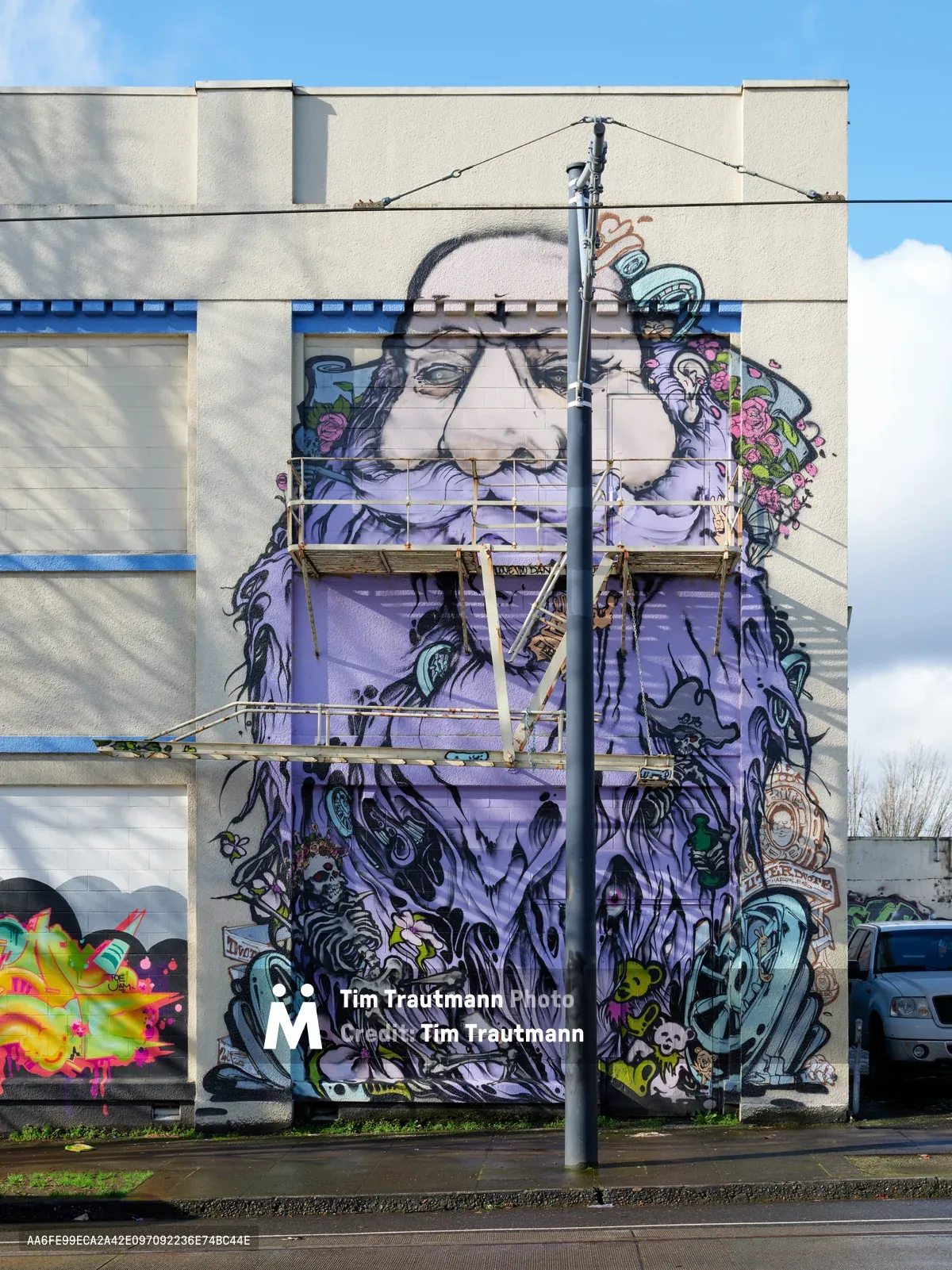 A monumental street mural dominates the side wall of a former Salvation Army warehouse on Southeast Ash Street in Portland's Central Eastside district. The artwork features a striking purple-robed figure with glasses and flowing beard, surrounded by intricate details including roses, skulls, and fantastical creatures that cascade down the building's facade. Industrial scaffolding bisects the composition vertically, while wispy clouds drift across the pale blue afternoon sky above the concrete structure.