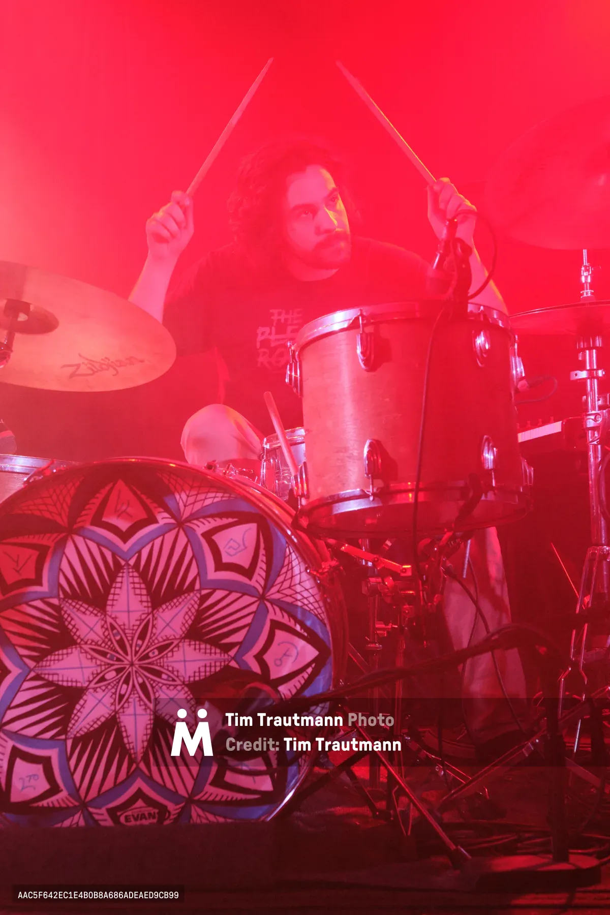 A drummer from Sun Blood Stories emerges through dense red stage lighting at Dante's nightclub in Portland's downtown district. The performer, captured mid-beat with drumsticks raised, appears almost spectral behind the thick crimson fog that transforms the intimate venue into a fever dream. An ornate mandala-patterned drum head anchors the foreground while the atmospheric haze swallows the drum kit's chrome hardware, creating an otherworldly concert experience that defines Portland's underground music scene.