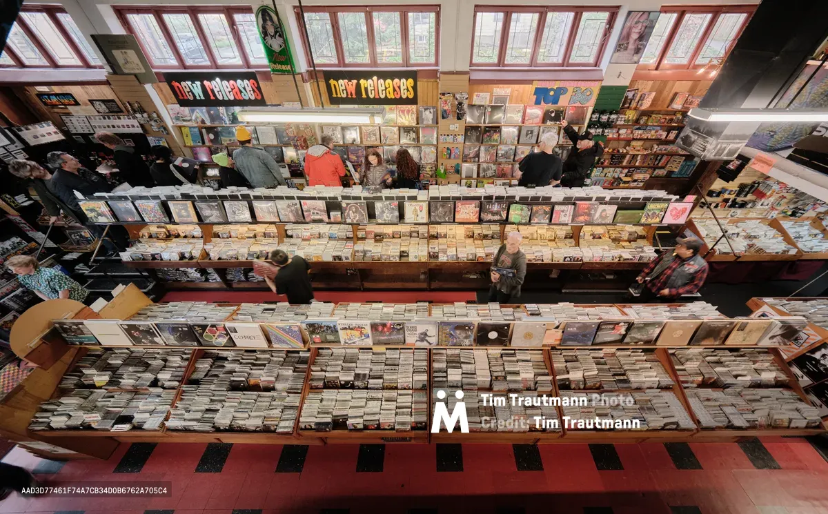 A commanding overhead perspective captures the bustling interior of Music Millennium during Record Store Day, Portland's iconic independent record store on East Burnside Street. Vinyl enthusiasts browse through meticulously organized wooden bins that stretch across multiple levels, their movements creating a dynamic human mosaic against the geometric patterns of thousands of album covers. Natural light streams through large windows, illuminating the "new releases" signage and casting warm shadows across the red and black striped flooring below.