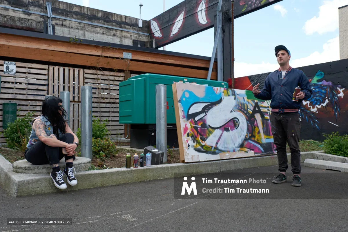 Galen Malcolm of the Portland Street Art Alliance demonstrates spray painting techniques at the Taylor Electric Blocks in Portland's Hosford-Abernethy neighborhood. A tattooed observer sits contemplatively on concrete steps while Malcolm stands beside his vibrant work in progress, paint cans scattered around the industrial setting. The scene captures the intersection of urban art culture and community education beneath overcast Pacific Northwest skies.