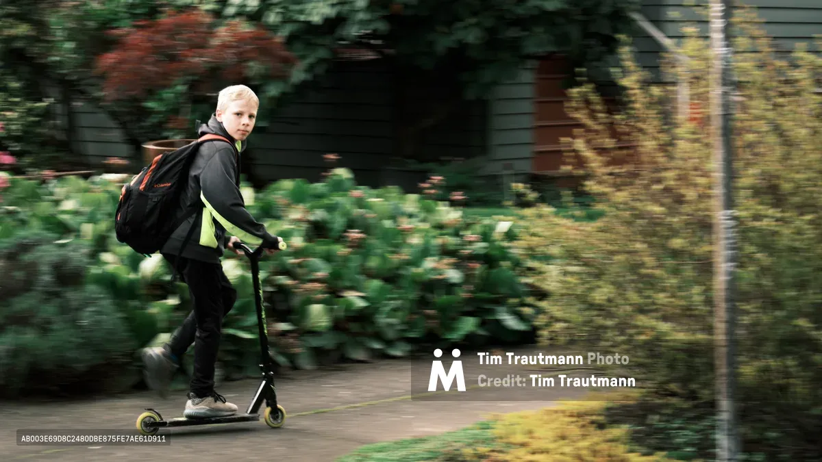 A blonde child in motion blur streaks through Portland's tree-lined Irvington district on a black scooter, their neon-green shirt creating a vivid focal point against the autumn foliage. The panning technique captures the kinetic energy of youth while rendering the surrounding residential landscape—weathered wood fences and mature maples—into impressionistic streaks of green and gold. The child's backward glance suggests both confidence and playful awareness as they navigate the quiet Northeast Portland street.