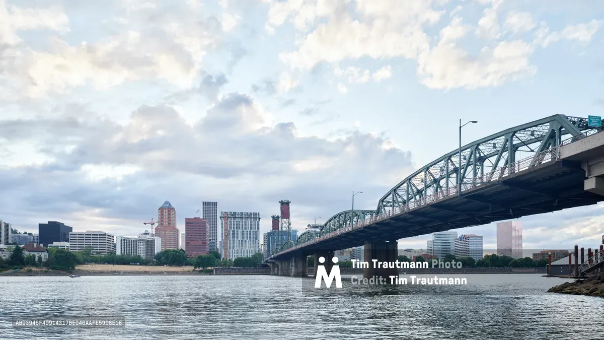 The iconic steel arch of the Hawthorne Bridge spans the rippling waters of the Willamette River, framing Portland's eclectic downtown skyline beneath a canvas of soft gray clouds. The city's mix of modern high-rises and historic brick buildings creates a compelling contrast against the industrial elegance of the green-painted bridge structure. Gentle afternoon light illuminates the scene with muted tones, while the river's surface reflects the urban landscape in shimmering fragments.