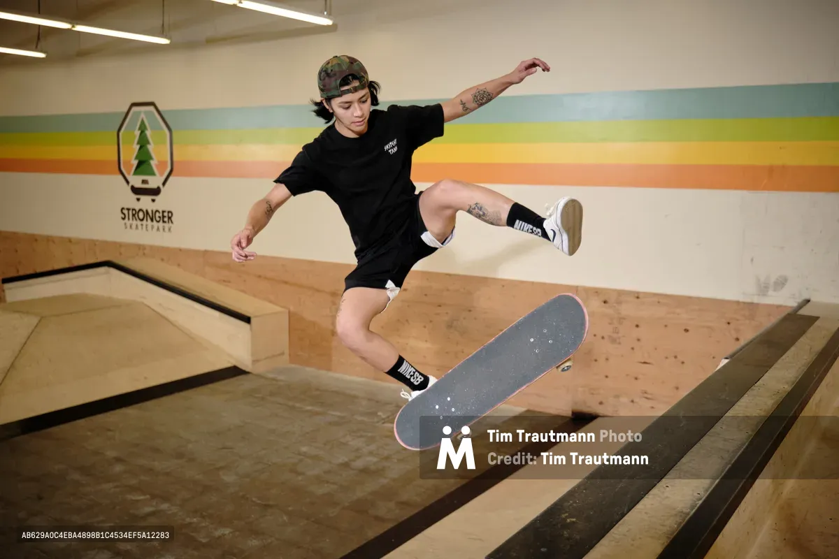 Professional skateboarder Christiana Means executes a precise aerial maneuver above the wooden bowl at Stronger Skatepark in Milwaukie, Oregon. Suspended mid-flight with arms extended for balance, she demonstrates complete control as her skateboard hovers beneath her. The indoor facility's warm lighting illuminates the rainbow-striped mural backdrop, creating a dynamic contrast against the natural wood construction of the skate bowl.