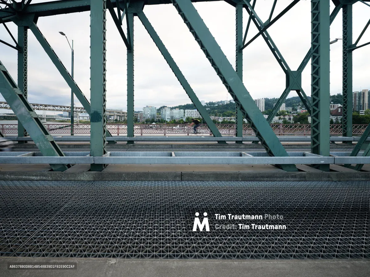 Industrial geometry frames the Portland skyline through the steel lattice work of the historic Hawthorne Bridge. Weathered green trusses create a rhythmic pattern of triangular supports, their riveted joints speaking to early 20th century engineering. Beyond the bridge's skeletal framework, modern residential towers rise against forested hills under an overcast Pacific Northwest sky, while the textured metal grating beneath adds tactile depth to this architectural intersection of past and present.