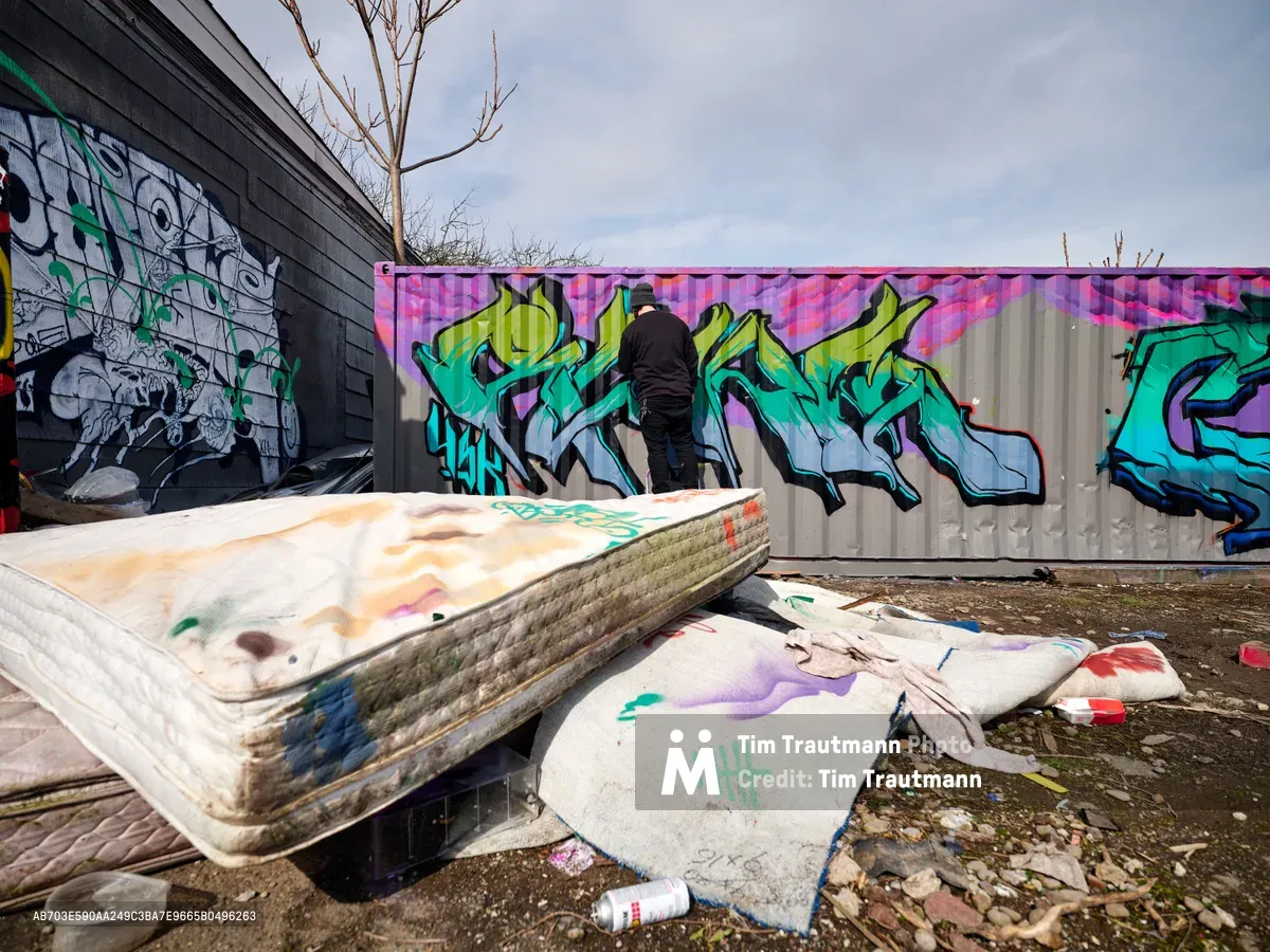 A street artist in dark clothing works intently on a colorful graffiti mural covering a white wall in Northeast Portland, Oregon. The scene captures the creative process amid an urban landscape of discarded mattresses, spray paint debris, and existing street art on nearby brick walls. Overcast skies and bare winter branches frame this raw documentation of underground art culture, while vibrant greens, purples, and blues emerge from the artist's aerosol work.