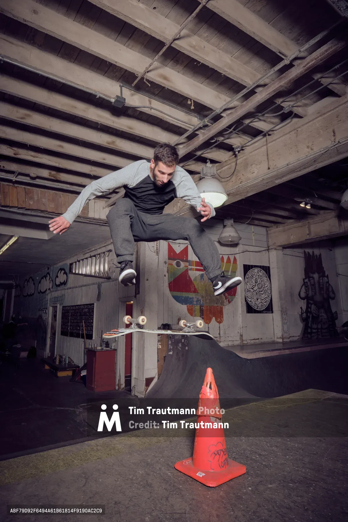 A skateboarder launches into the air above a bright orange traffic cone in the dimly lit Commonwealth Skateboarding in Portland's Buckman neighborhood. The raw industrial space features exposed wooden ceiling beams, graffiti-covered walls, and makeshift ramps, creating an authentic underground skate culture atmosphere. Moody lighting from overhead fixtures casts dramatic shadows across the concrete floor, while colorful street art and skateboard graphics provide vibrant contrast against the weathered interior.