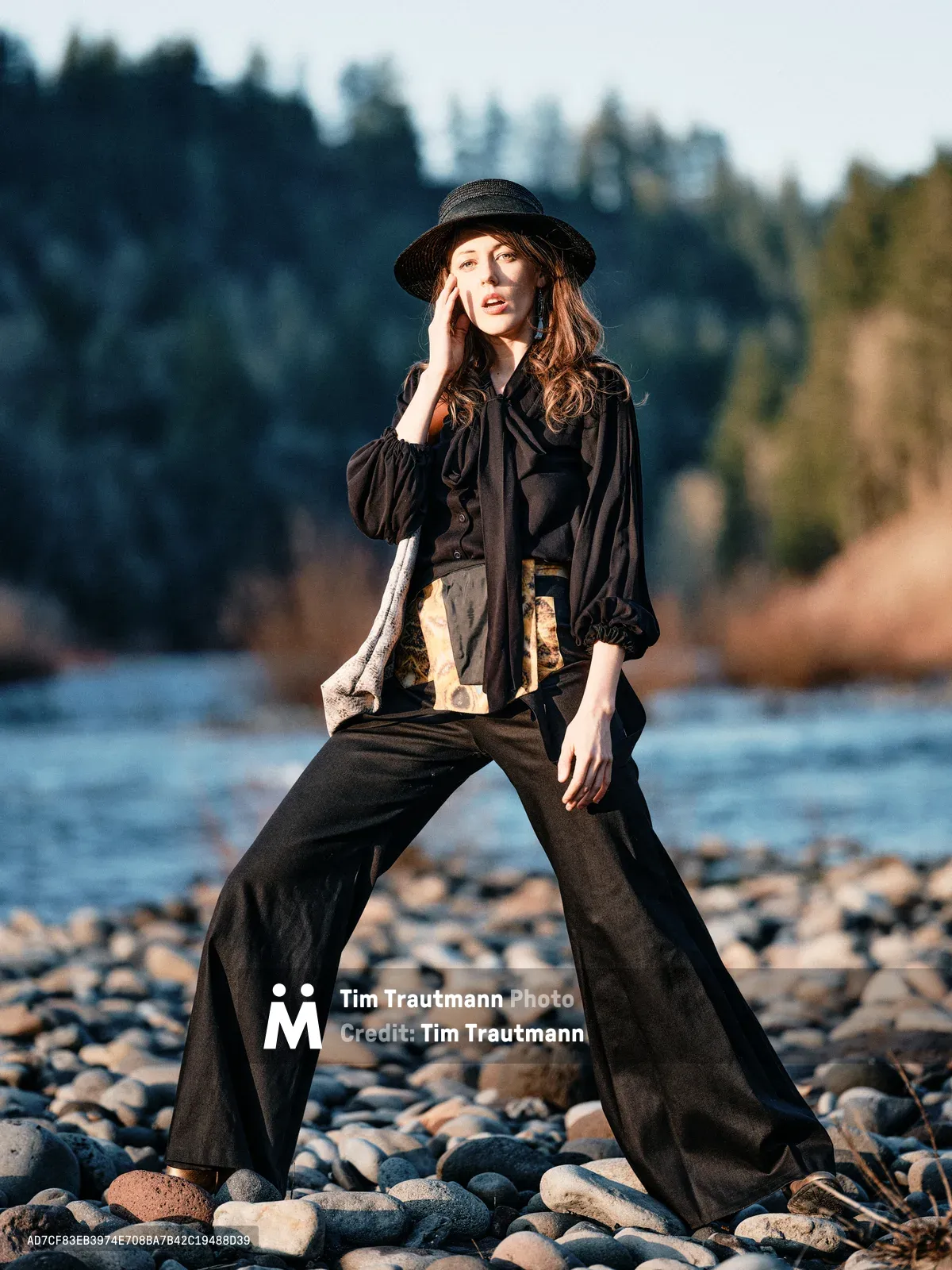 A woman in flowing black attire and wide-brimmed hat stands contemplatively on the rocky shoreline of the Columbia River along Multnomah County's Floodplain Trail. The golden hour light illuminates her thoughtful pose as she raises one hand to her temple, her wide-leg trousers and billowing blouse creating dramatic silhouettes against the water. The background reveals the characteristic forested hills of the Columbia River Gorge, with smooth river stones scattered beneath her feet on the exposed riverbed.