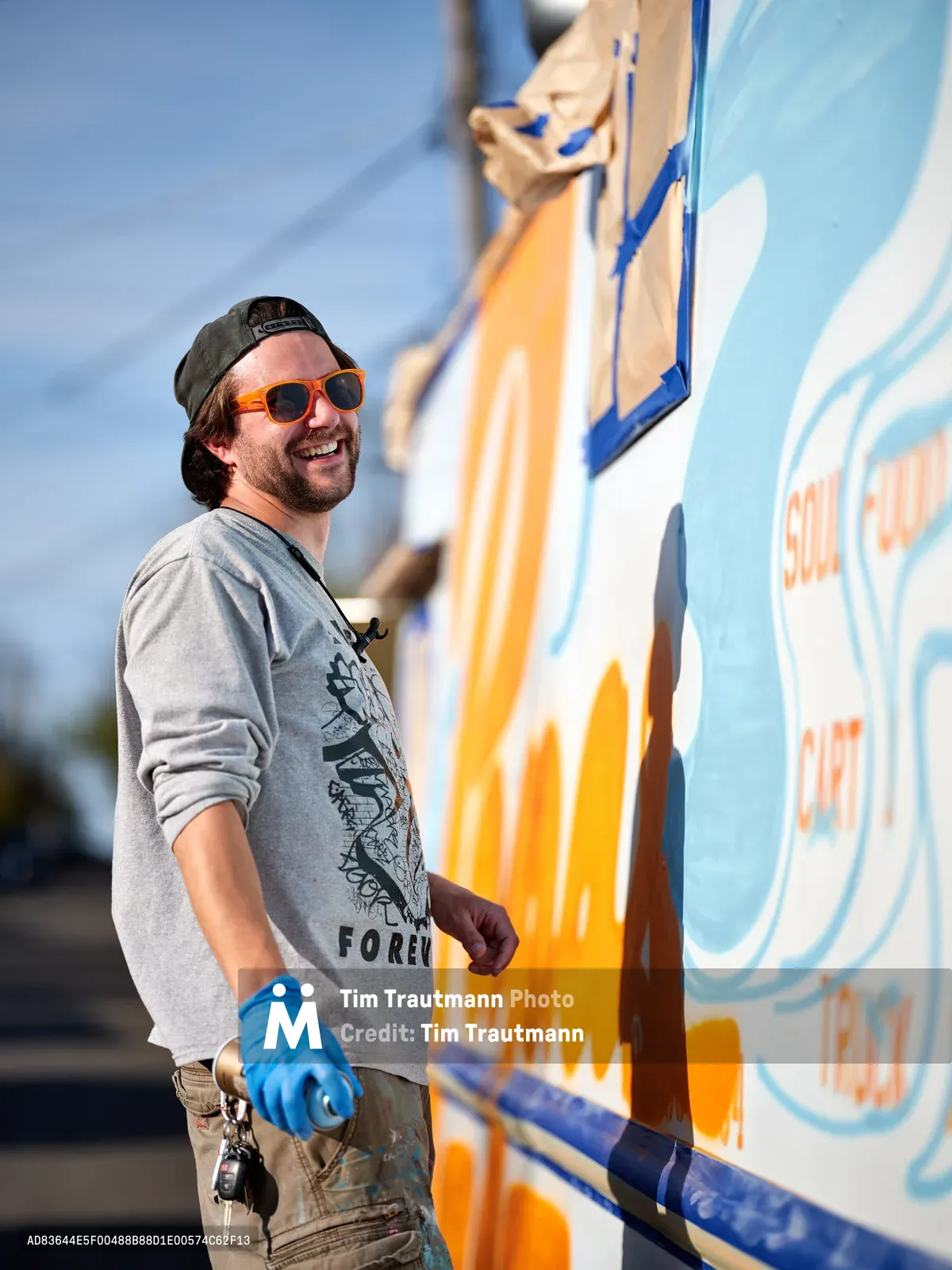 A bearded muralist in orange sunglasses and a backwards cap grins while working on a colorful food truck exterior in Portland's King neighborhood. Wearing blue protective gloves and a gray graphic sweatshirt, he stands before his vivid orange and blue artwork that transforms the vehicle's surface. The bright afternoon sunlight illuminates both the artist and his dynamic street art, capturing the creative energy of Portland's thriving food truck culture.