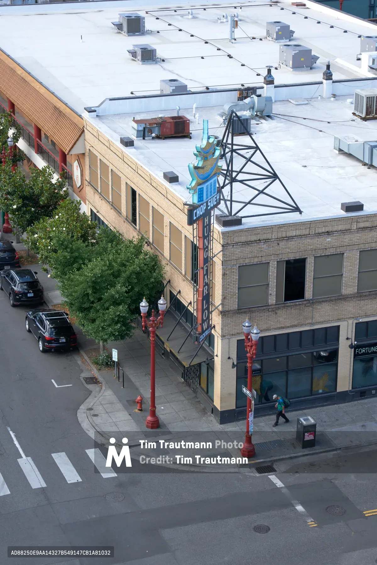 An elevated perspective captures the intersection where traditional meets contemporary in Portland's Chinatown district. The geometric precision of white crosswalk stripes guides the eye through a scene where vintage brick architecture houses modern commerce, while HVAC units dot the pristine white rooftop like mechanical sentinels. Red street lamps stand as colorful punctuation marks against the muted palette of concrete and brick, their warm hue echoing the neighborhood's cultural heritage.