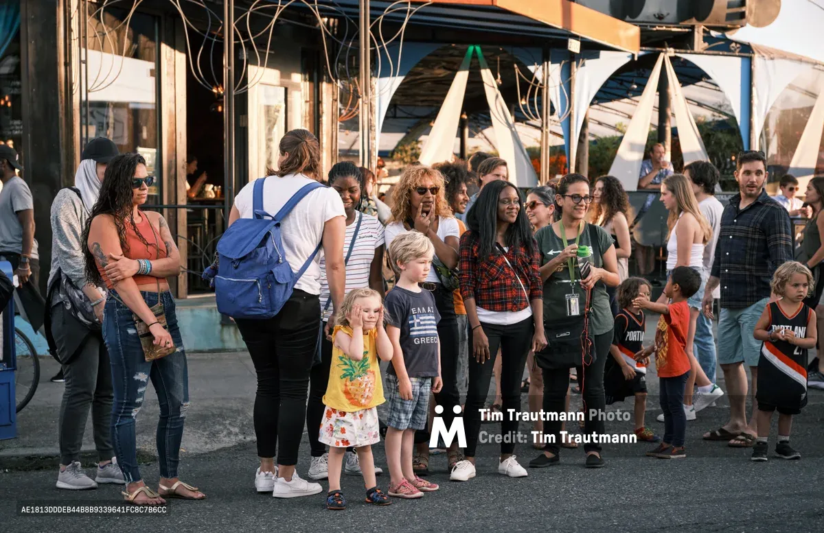 Families and friends congregate along Northeast Alberta Street during Portland's Last Thursday street fair, bathed in warm evening light. The diverse crowd spans generations, from toddlers in colorful summer clothing to adults carrying blue backpacks and wearing casual attire. Behind them, the characteristic storefronts and white fabric installations create an urban festival backdrop, while the golden hour sun casts long shadows across the asphalt.