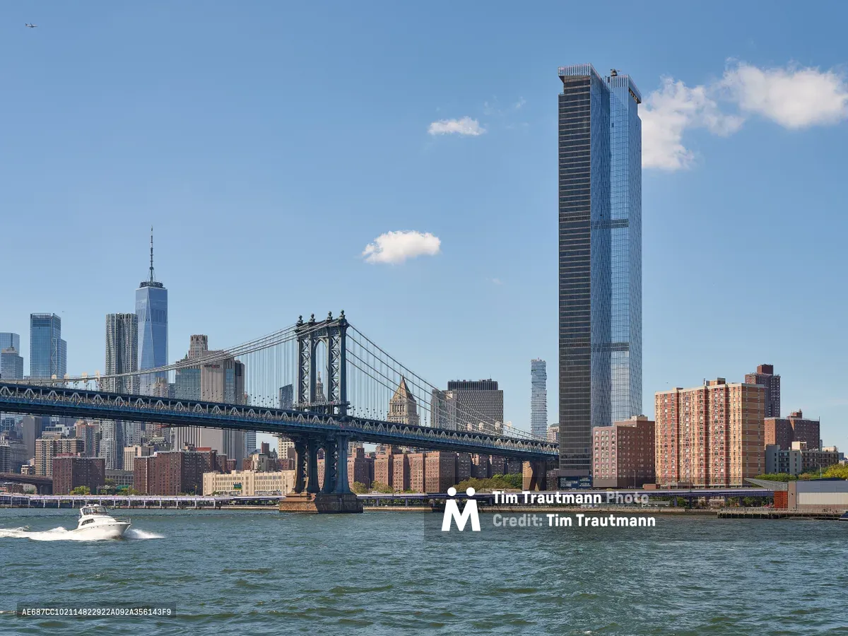 The iconic Manhattan Bridge stretches across the East River in elegant suspension, its steel towers and cables creating geometric lines against a pale blue afternoon sky dotted with cotton-white clouds. One World Trade Center's gleaming spire punctuates the left side of Manhattan's financial district, while a striking dark glass tower dominates the right portion of the frame. A white pleasure boat cuts through the choppy river waters in the foreground, leaving a gentle wake as it passes beneath this architectural symphony of old and new New York.