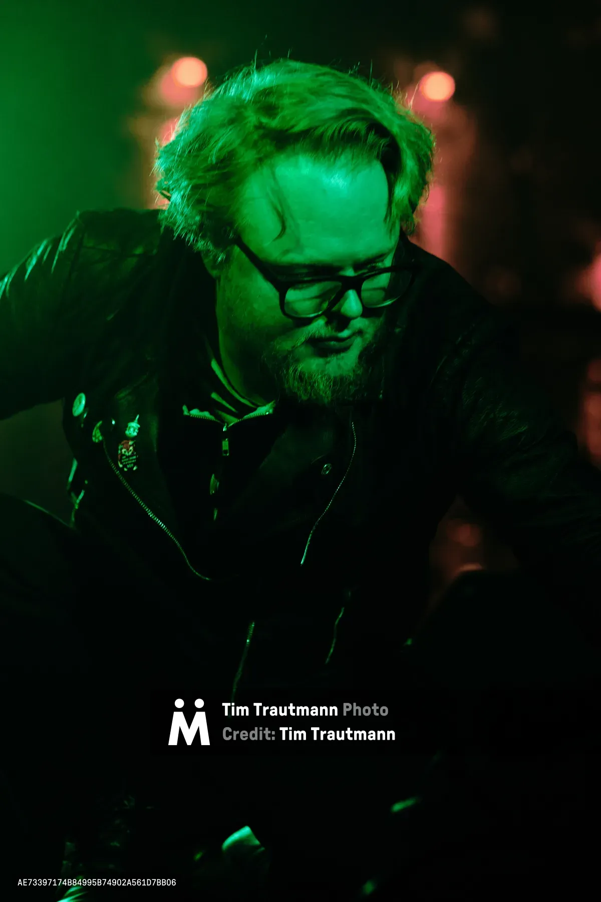 Electronic musician Eli Savage of Ellipsism performs under intense emerald stage lighting at Dante's in Portland's downtown district. The atmospheric green wash creates a cyberpunk aesthetic, casting the bearded artist in an otherworldly glow as he concentrates over his equipment. Warm bokeh lights pierce through the darkness behind him, while his leather jacket and glasses catch the saturated color, creating a moody portrait of underground electronic music culture.