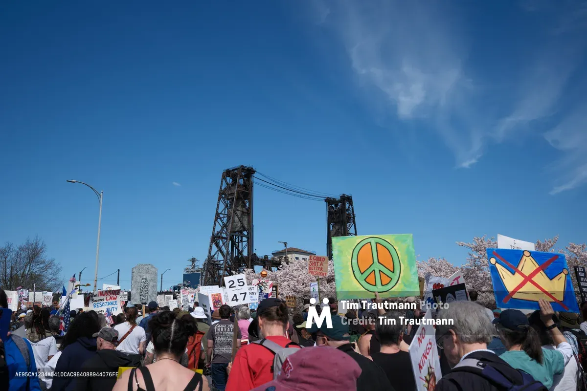 Hundreds of demonstrators gather beneath Portland's iconic Steel Bridge on Northwest Naito Parkway, their colorful protest signs creating a vibrant tapestry against the industrial backdrop. A prominent green peace symbol banner cuts through the crowd while cherry blossoms frame the scene, their delicate pink petals contrasting with the raw steel lattework above. The crystalline blue sky and brilliant spring light illuminate this grassroots political action, capturing the intersection of urban infrastructure and democratic expression in Oregon's largest city.