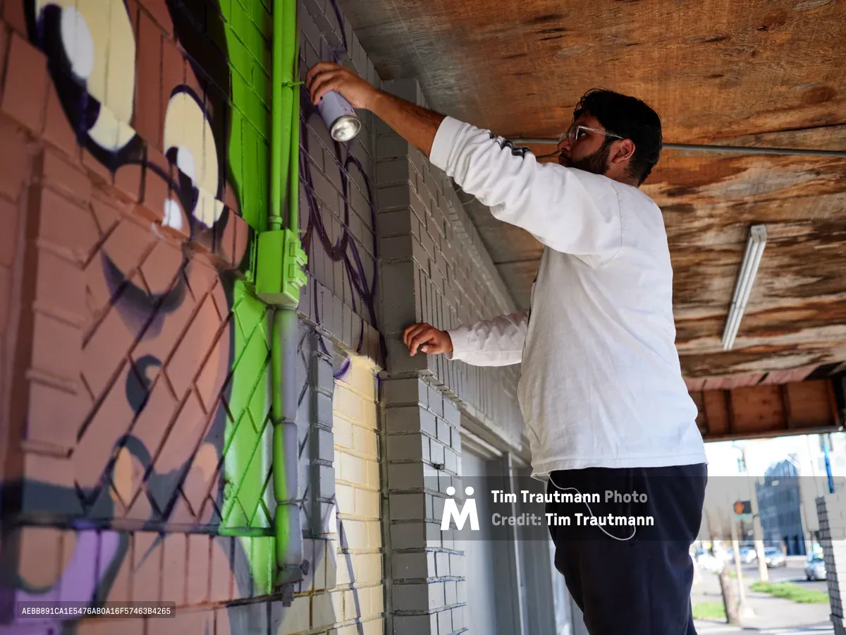 Muralist Heysus extends his reach toward weathered brick walls beneath the exposed wooden beams of a Northeast Portland storefront. Spray paint can in hand, he adds vibrant purple and green strokes to an emerging street art composition, his white hoodie catching the filtered daylight that streams through this covered walkway. The urban canvas stretches across textured surfaces where old meets new in Portland's evolving Alberta Arts District.
