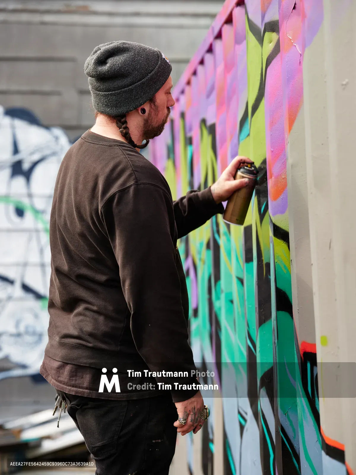 A bearded artist in a charcoal beanie and brown hoodie concentrates intently as he applies spray paint to a colorful mural on a concrete wall in Northeast Portland, Oregon. The artwork features flowing organic shapes in brilliant pinks, greens, and yellows that contrast sharply against the gray urban surface. Soft natural light illuminates the scene, capturing the focused energy of creative expression in an industrial setting. Additional graffiti work is visible in the background, establishing this as part of Northeast Portland's thriving street art scene.