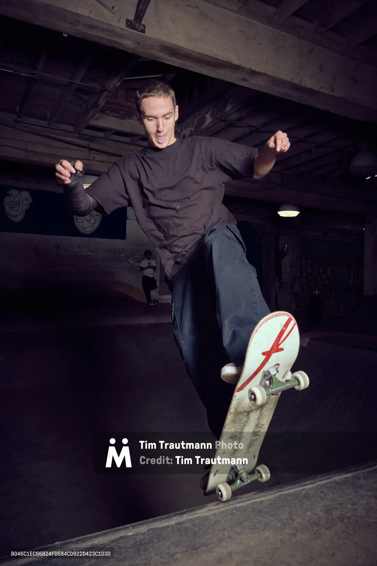 A skateboarder executes a fluid maneuver in the shadowy concrete bowl at Commonwealth Skateboarding in Portland's Buckman neighborhood. Dressed in a dark sweatshirt and loose jeans, the rider navigates the curved transition with focused intensity, his weathered deck bearing a distinctive red lightning bolt graphic. The underground skate park's exposed concrete beams and industrial lighting create a gritty urban cathedral for this moment of athletic grace.