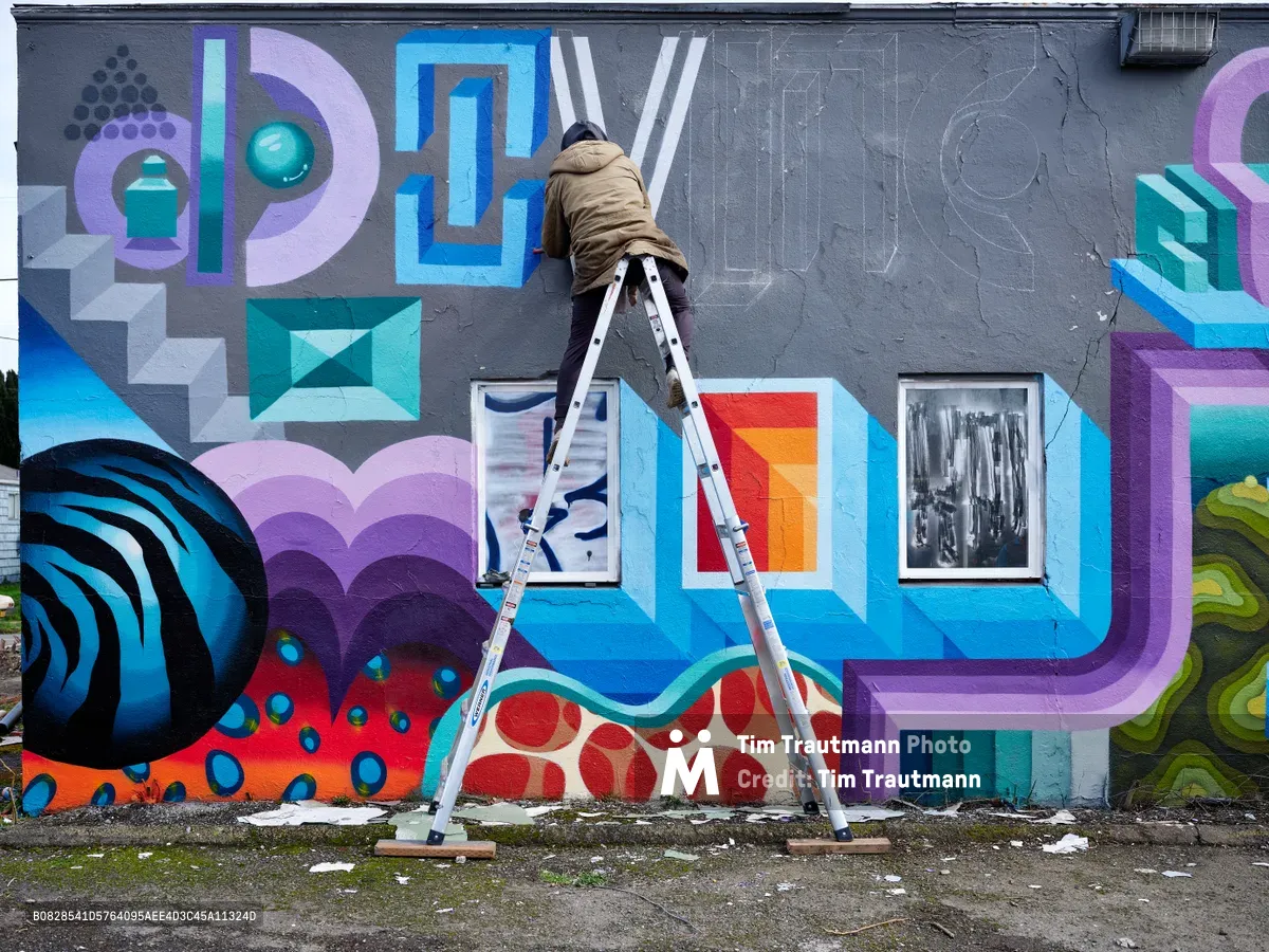 A hooded artist perched on an aluminum ladder applies paint to a kaleidoscopic mural sprawling across a gray concrete wall in Northeast Portland, Oregon. The dynamic composition bursts with geometric shapes, dimensional letterforms, and embedded photographic elements in vivid purples, blues, and oranges. Weathered pavement and urban debris frame the base of this evolving street art masterpiece, capturing the raw creative energy of the Pacific Northwest's renowned mural scene.