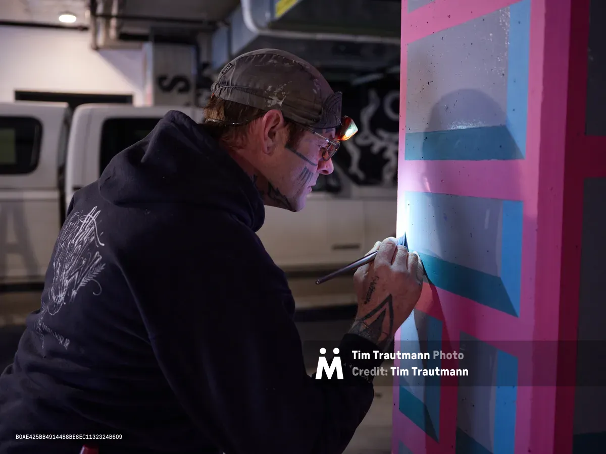 Portland artist and bike activist Rabbitt meticulously applies paint to a vibrant geometric mural in the dimly lit parking garage of the Clay Creative building in the Hosford-Abernethy neighborhood. Wearing a camo cap and dark hoodie, the tattooed artist works with focused precision on bold pink, blue, and purple angular forms that transform the industrial concrete space. The fluorescent lighting creates dramatic shadows as he brings color and artistry to the urban underground environment.