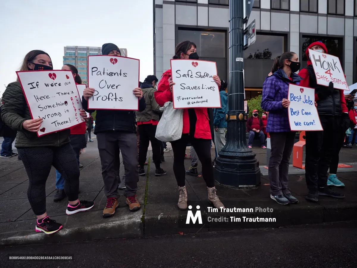 Healthcare workers gather on a grey Portland morning outside the Kaiser Permanente Tower in the Lloyd District, their handmade protest signs creating stark white rectangles against the building's glass facade. Masked nurses in winter jackets hold placards declaring "Patients Over Profits" and "Safe Staffing Saves Lives," their determination evident despite the overcast sky. The urban sidewalk scene captures the tension between corporate healthcare and frontline workers, with the tower's reflective windows looming behind the small but resolute group of demonstrators.