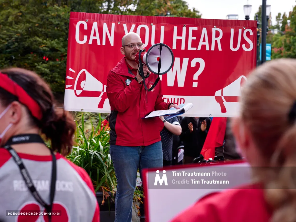 A bald man in a vibrant red jacket commands attention as he speaks through a megaphone at a Kaiser Permanente nurses' strike rally in Portland's Lloyd District. Behind him, a bold red and white banner poses the rhetorical question 'CAN YOU HEAR US NOW?' - its message emphasized by megaphone graphics. The speaker holds papers in one hand while addressing a crowd of supporters wearing matching red and white strike colors, creating a sea of solidarity that fills the foreground with purposeful energy.